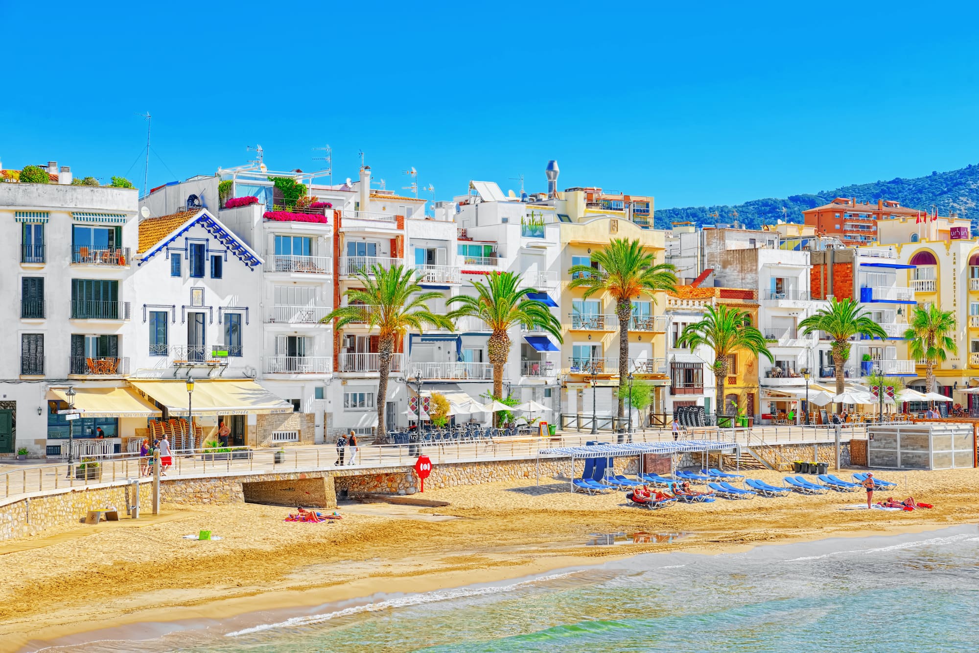 Sitges, España: casas coloridas frente al mar, playa de arena y palmeras.