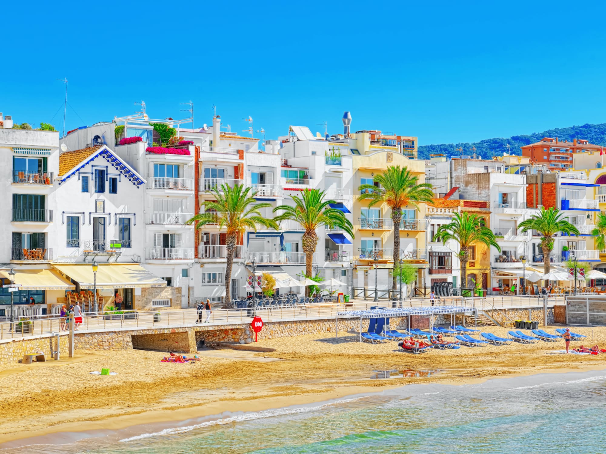 Colorful coastal town with white buildings, palm trees, sandy beach, and blue ocean.