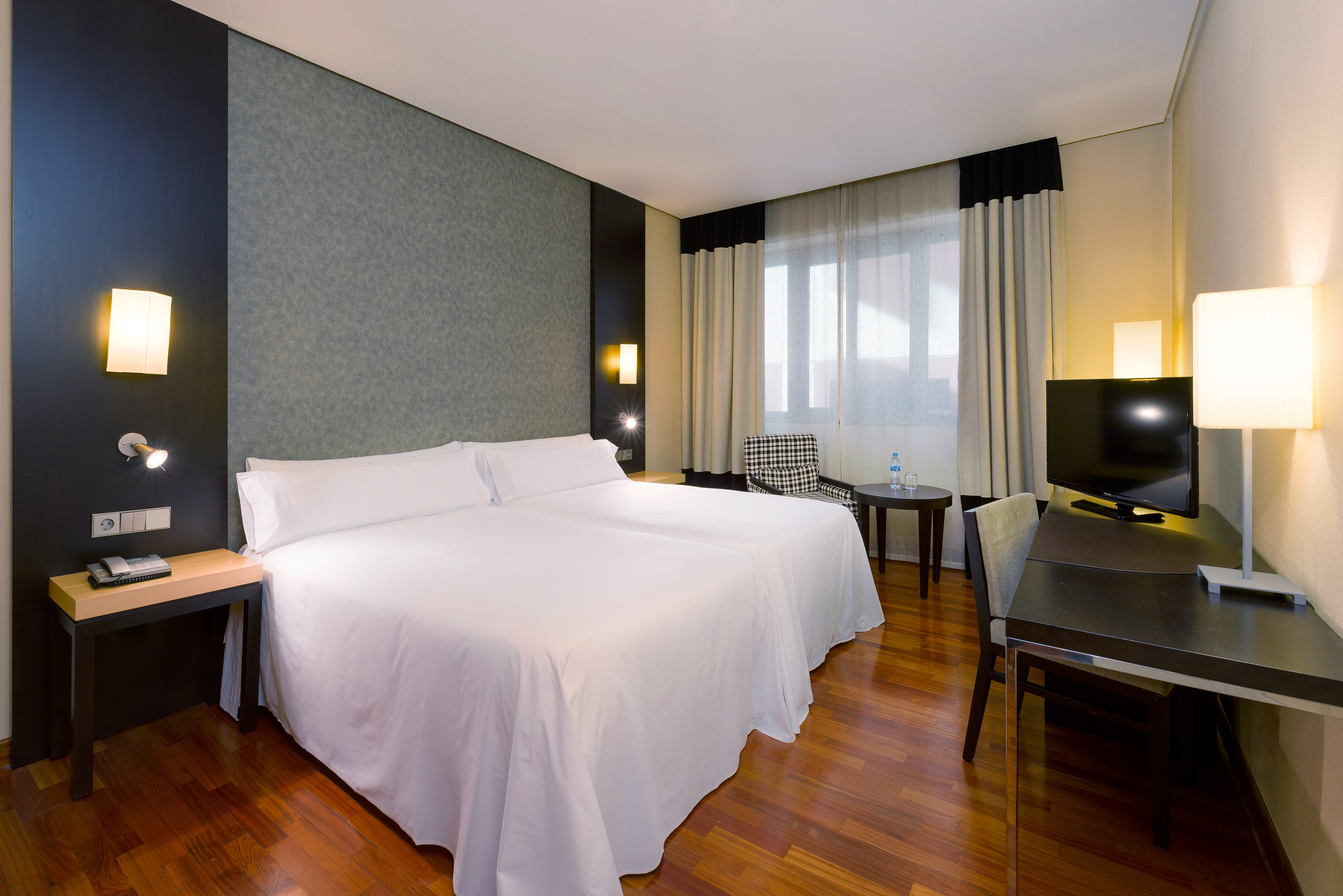 Modern hotel room with twin beds, wood floor, desk, TV, and window.