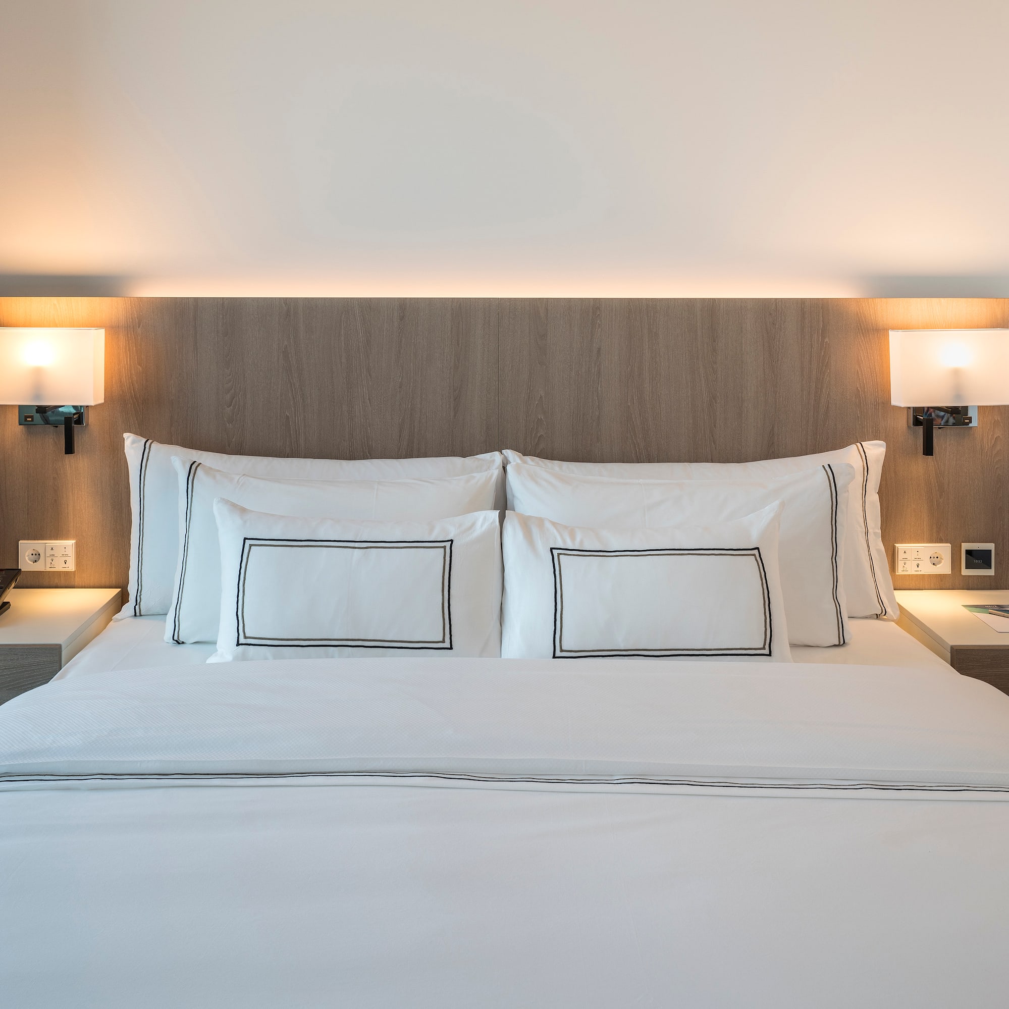 Modern hotel bed with white bedding, wooden headboard, and illuminated lamps.