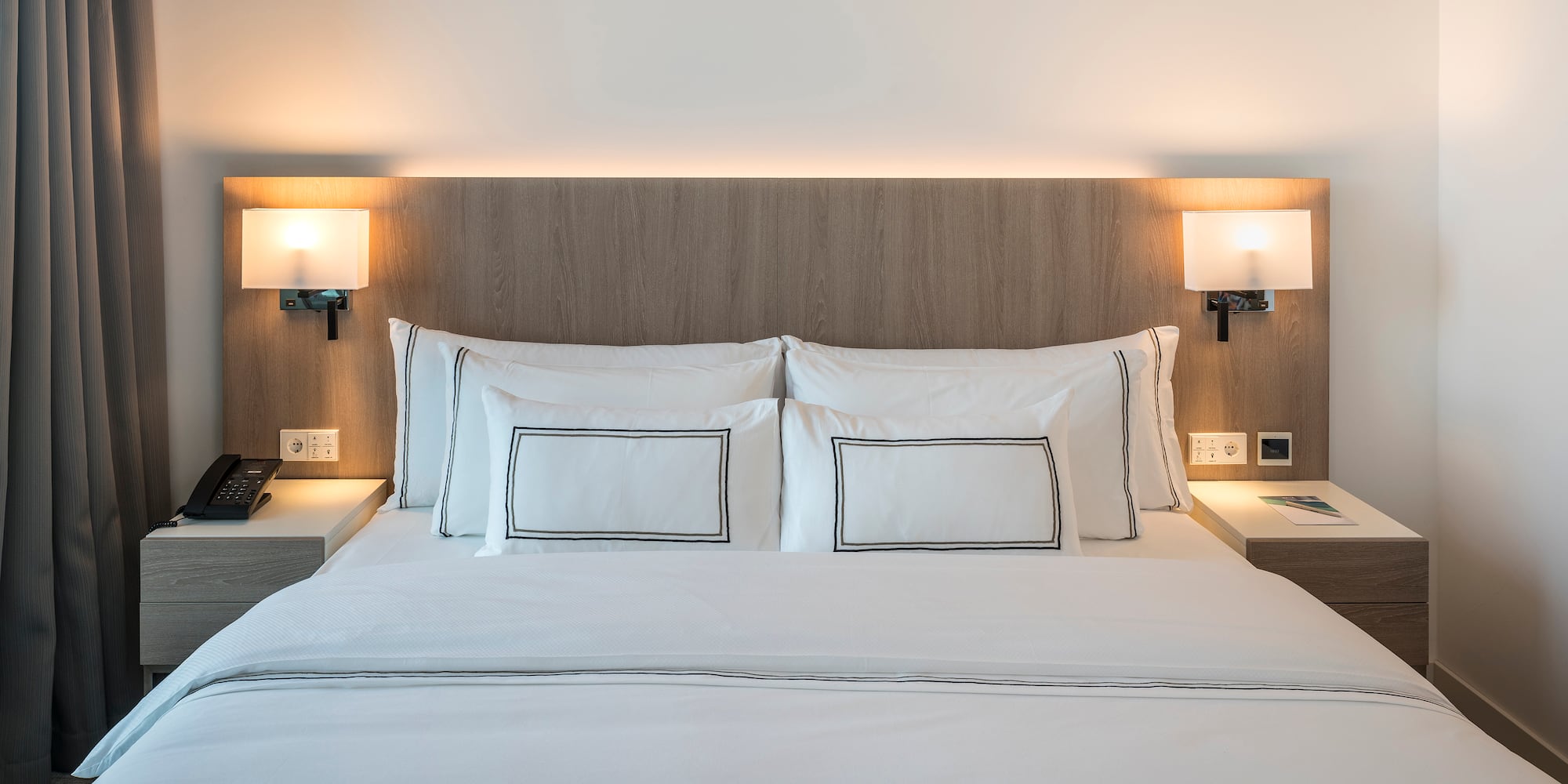 Modern hotel bed with white bedding, wooden headboard, and illuminated lamps.