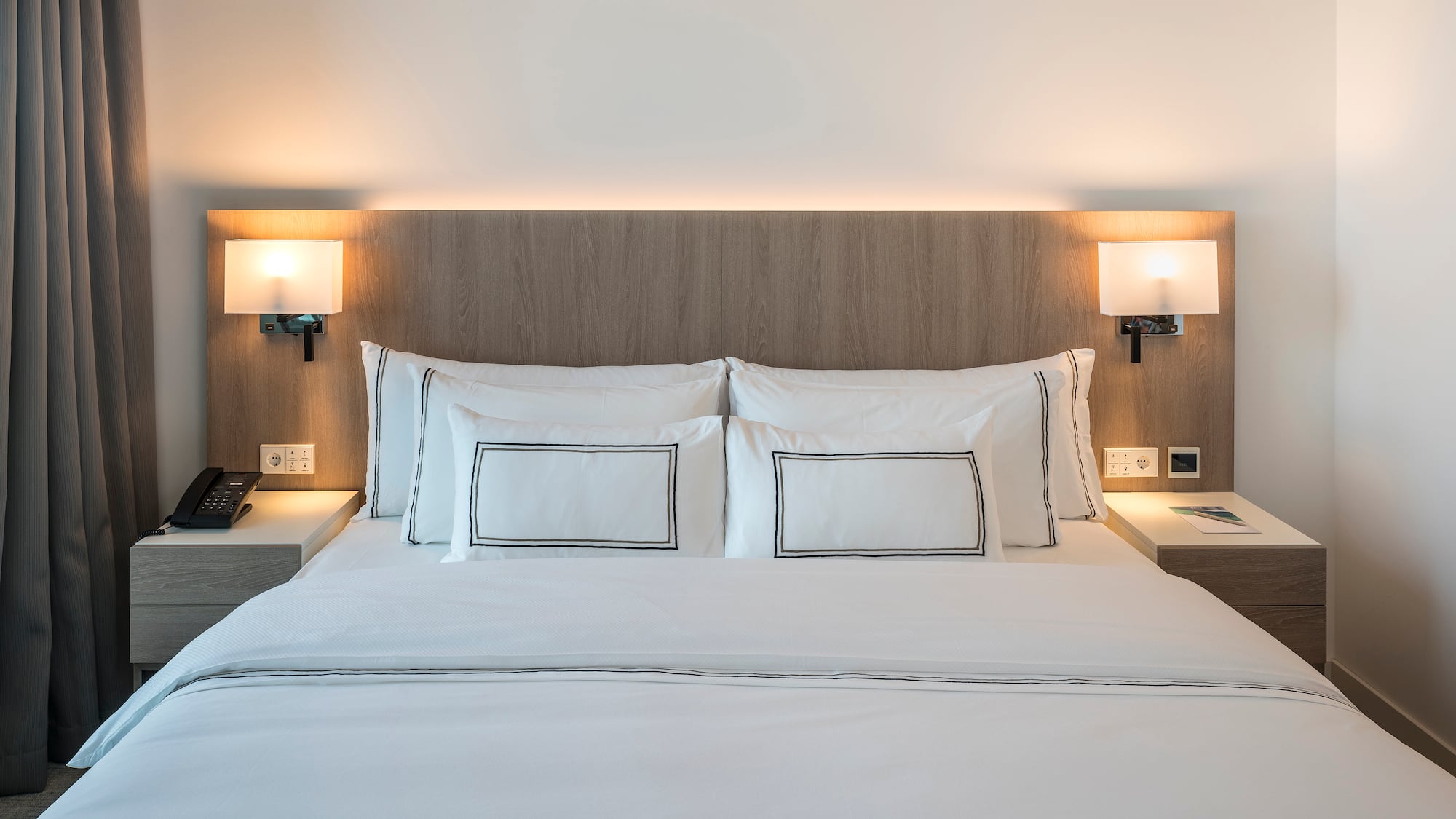 Modern hotel bed with white bedding, wooden headboard, and illuminated lamps.