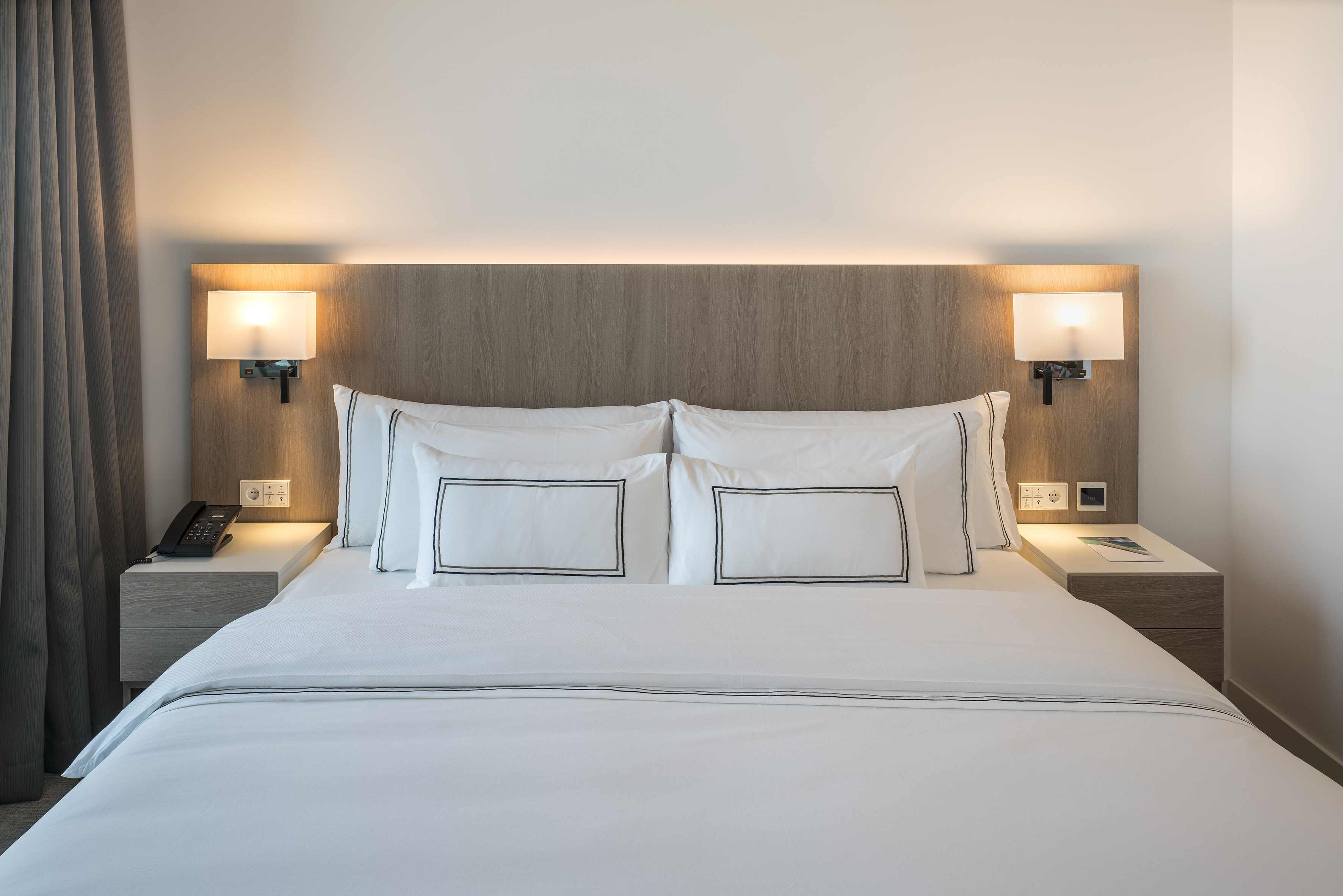 Modern hotel bed with white bedding, wooden headboard, and illuminated lamps.