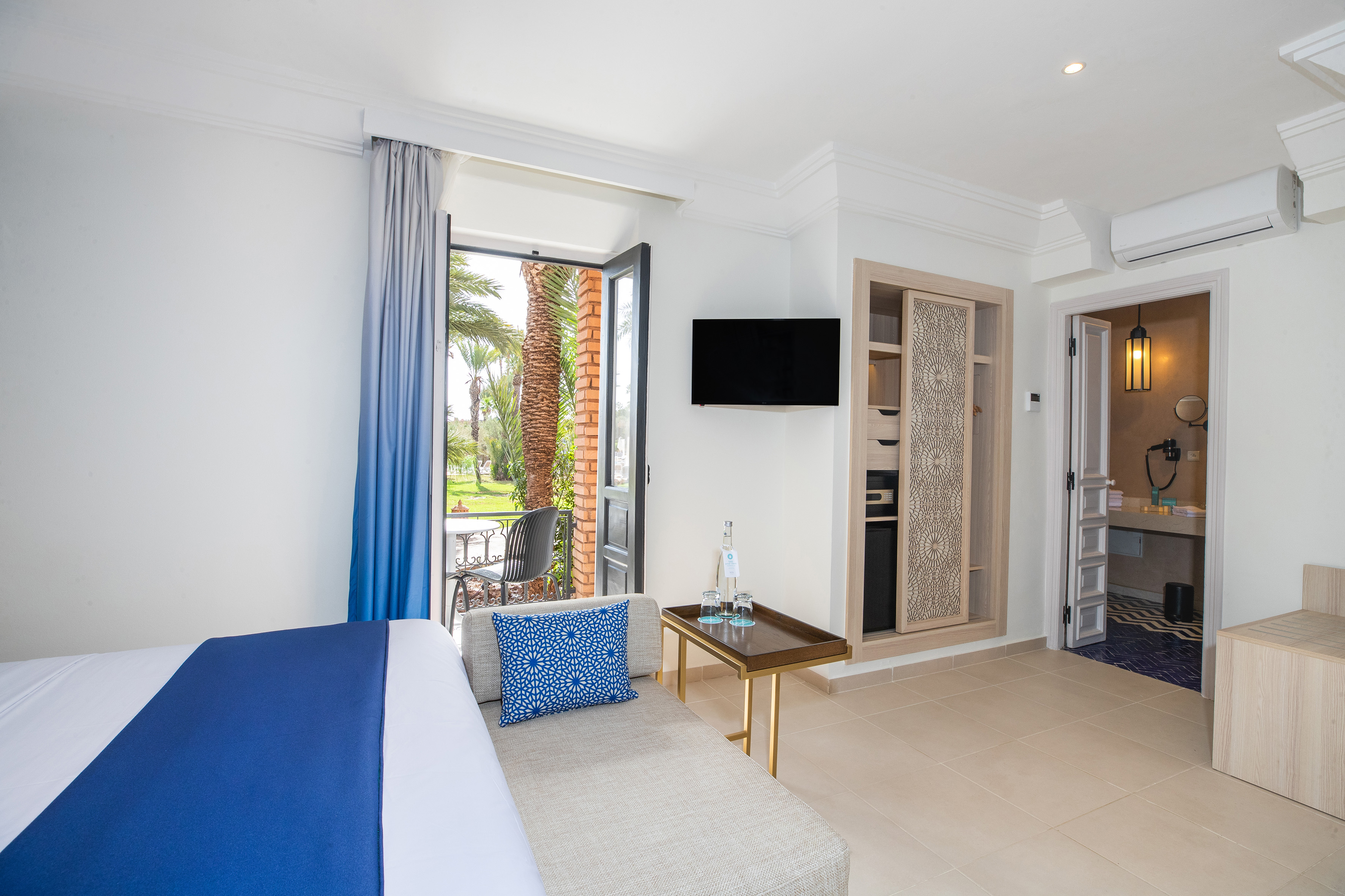 Hotel bedroom with balcony, palm garden view, TV, and decorative wardrobe.