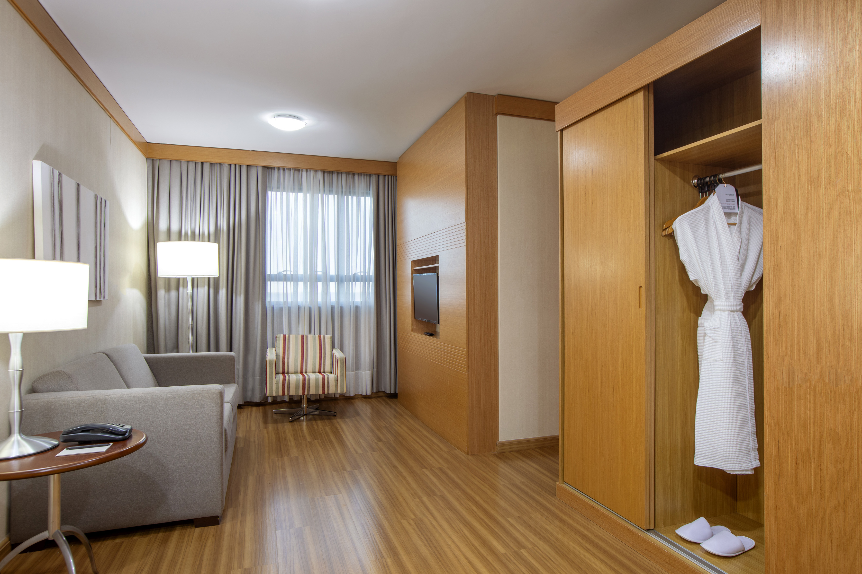 Modern hotel suite living area with sofa, TV, wooden wardrobe, and window.