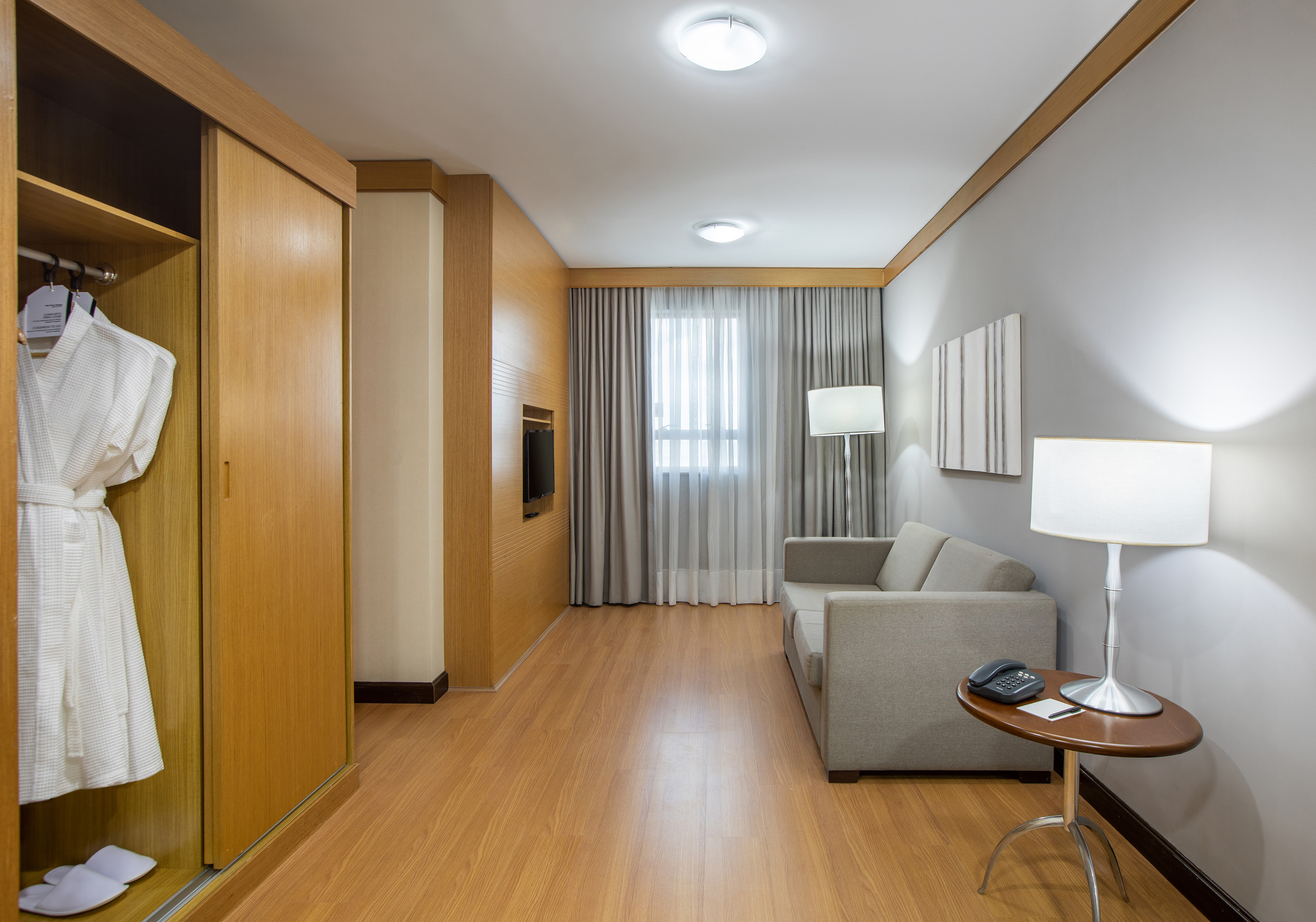 Modern hotel studio with wooden floor, sofa, wardrobe, and bright window.