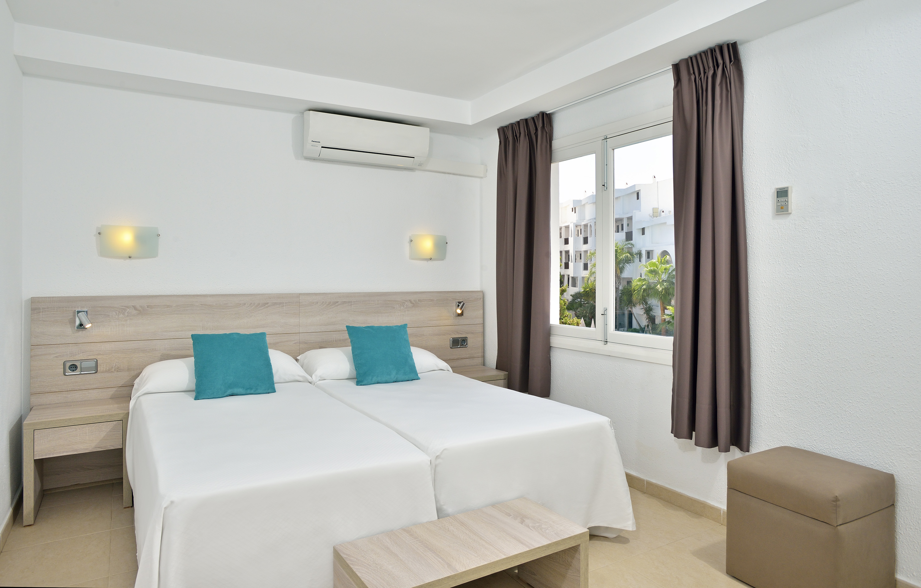 Bright hotel room with twin beds, white linens, teal pillows, AC, and window view.