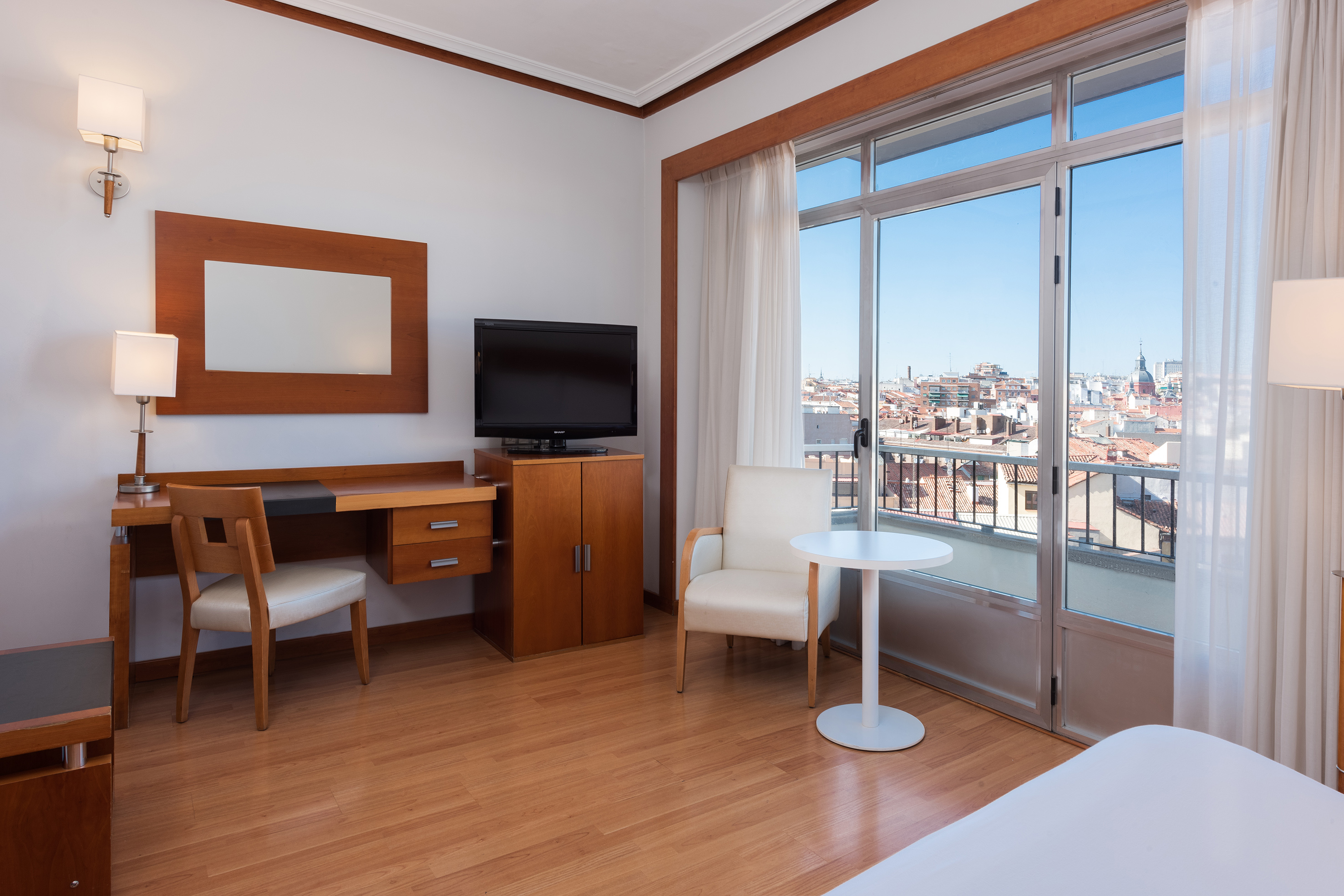 Modern hotel room with desk, TV, wood floor, and large window city view.