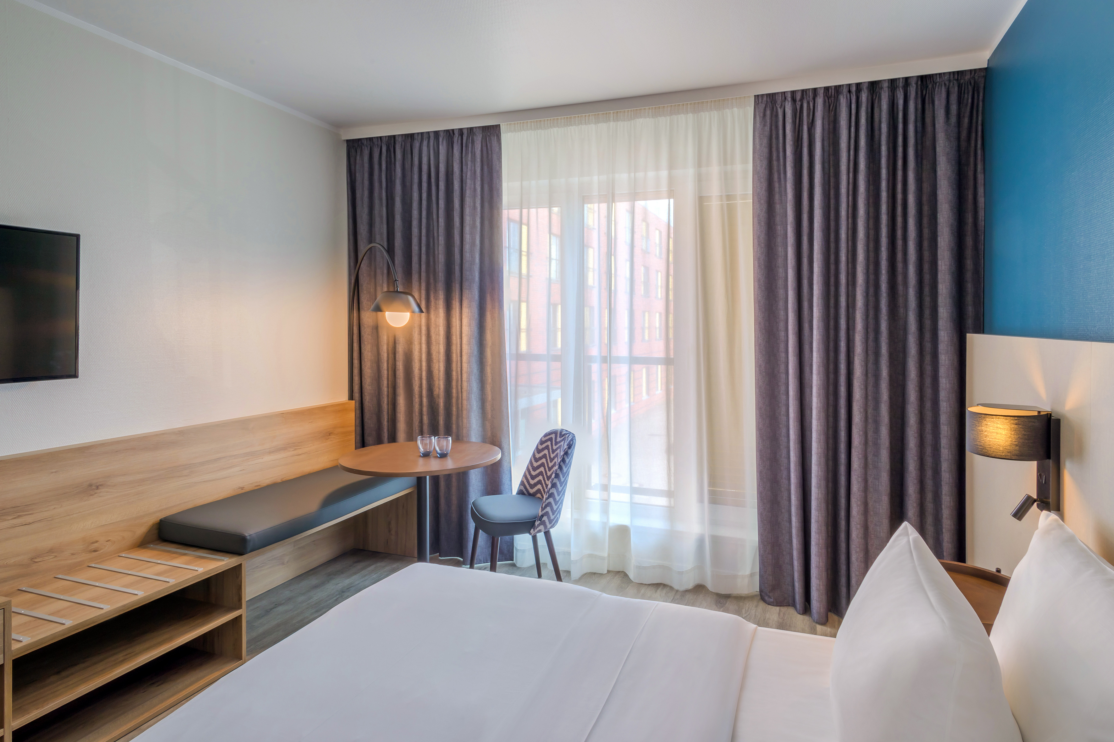 Modern hotel room with bed, wooden bench, table, and bright window.
