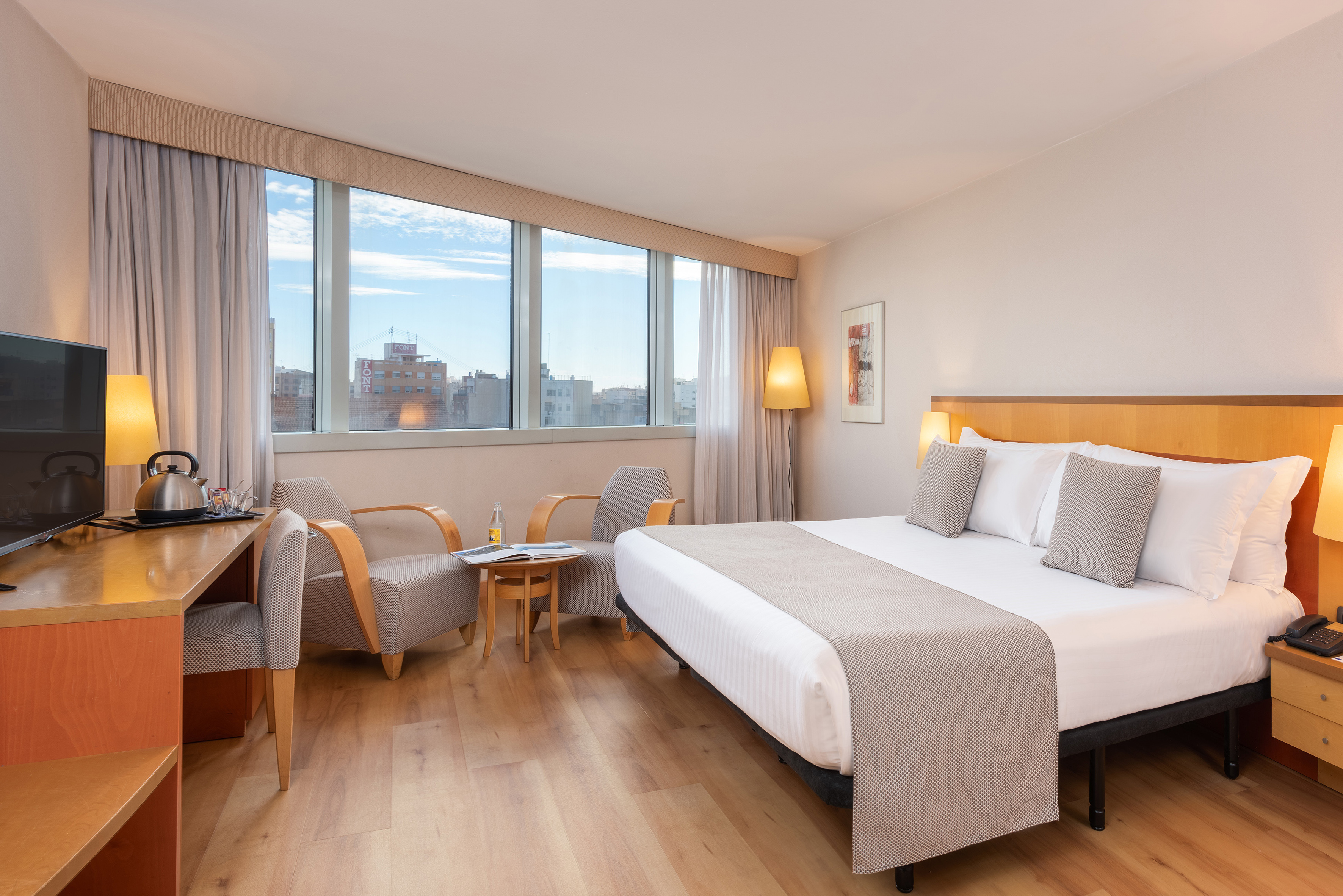 Bright hotel room with king bed, city view window, wooden floor, and desk.