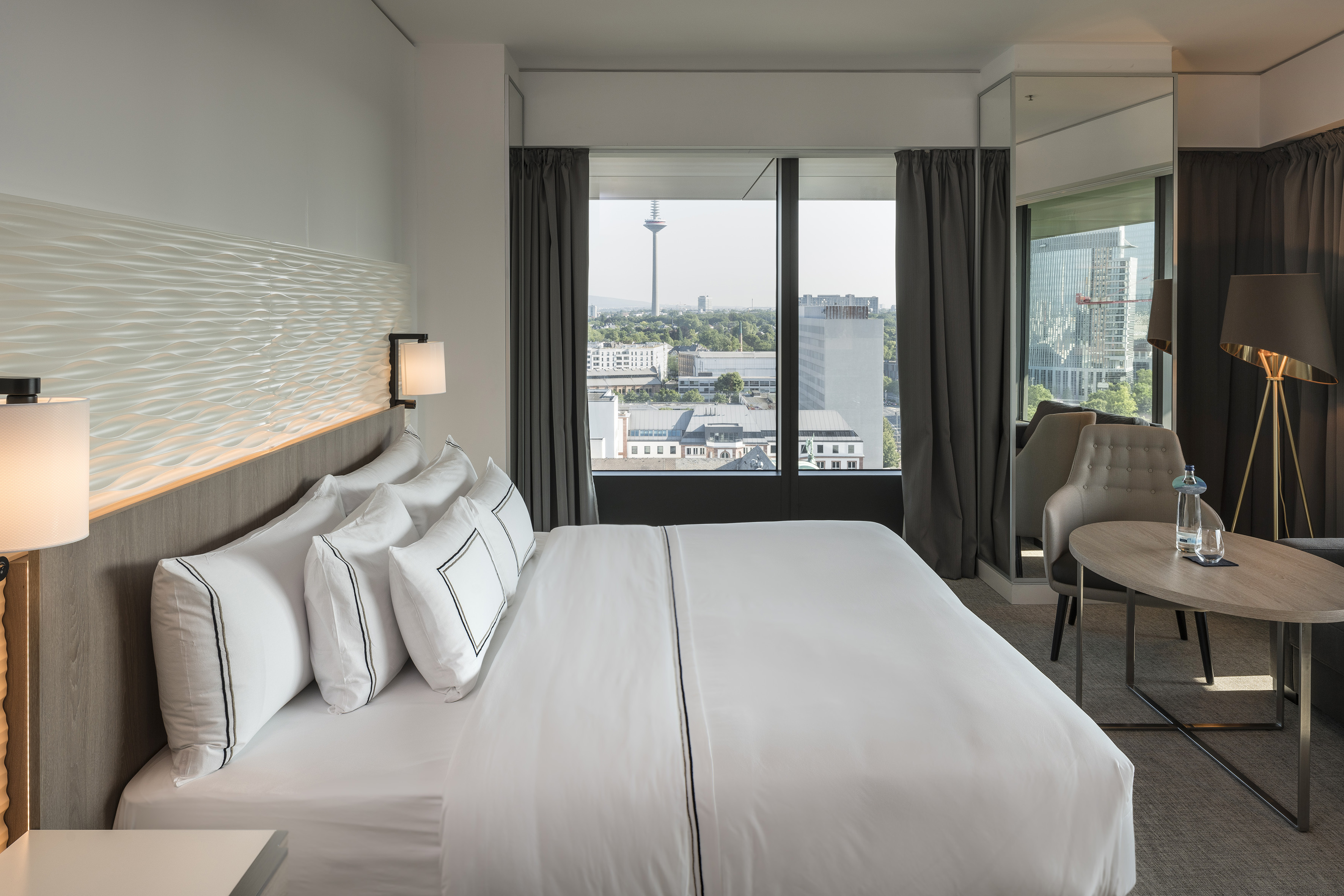 Modern hotel room with city view, Frankfurt Europaturm, and white bed.