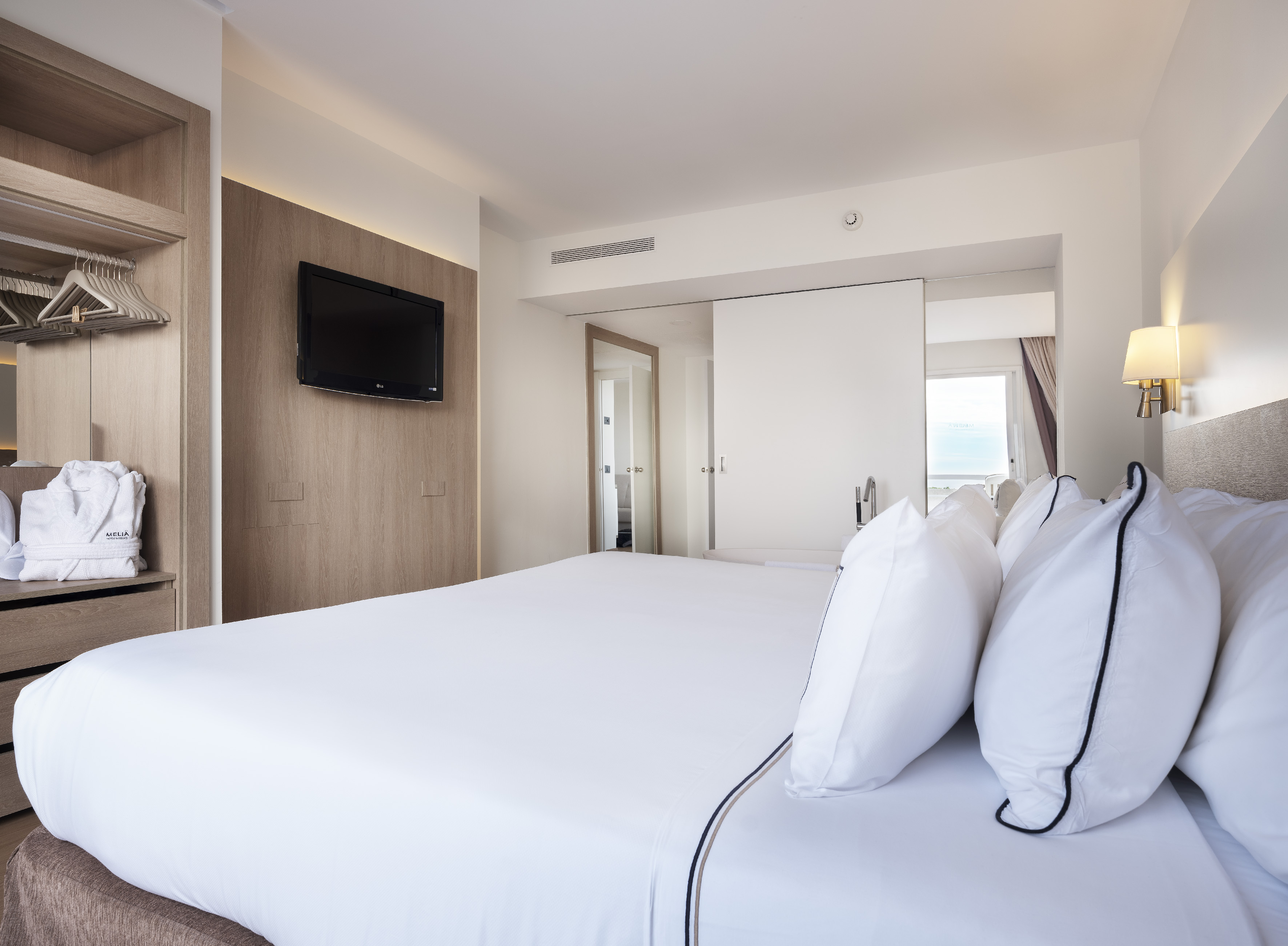 Bright hotel room with white bed, wall-mounted TV, and open wooden closet.