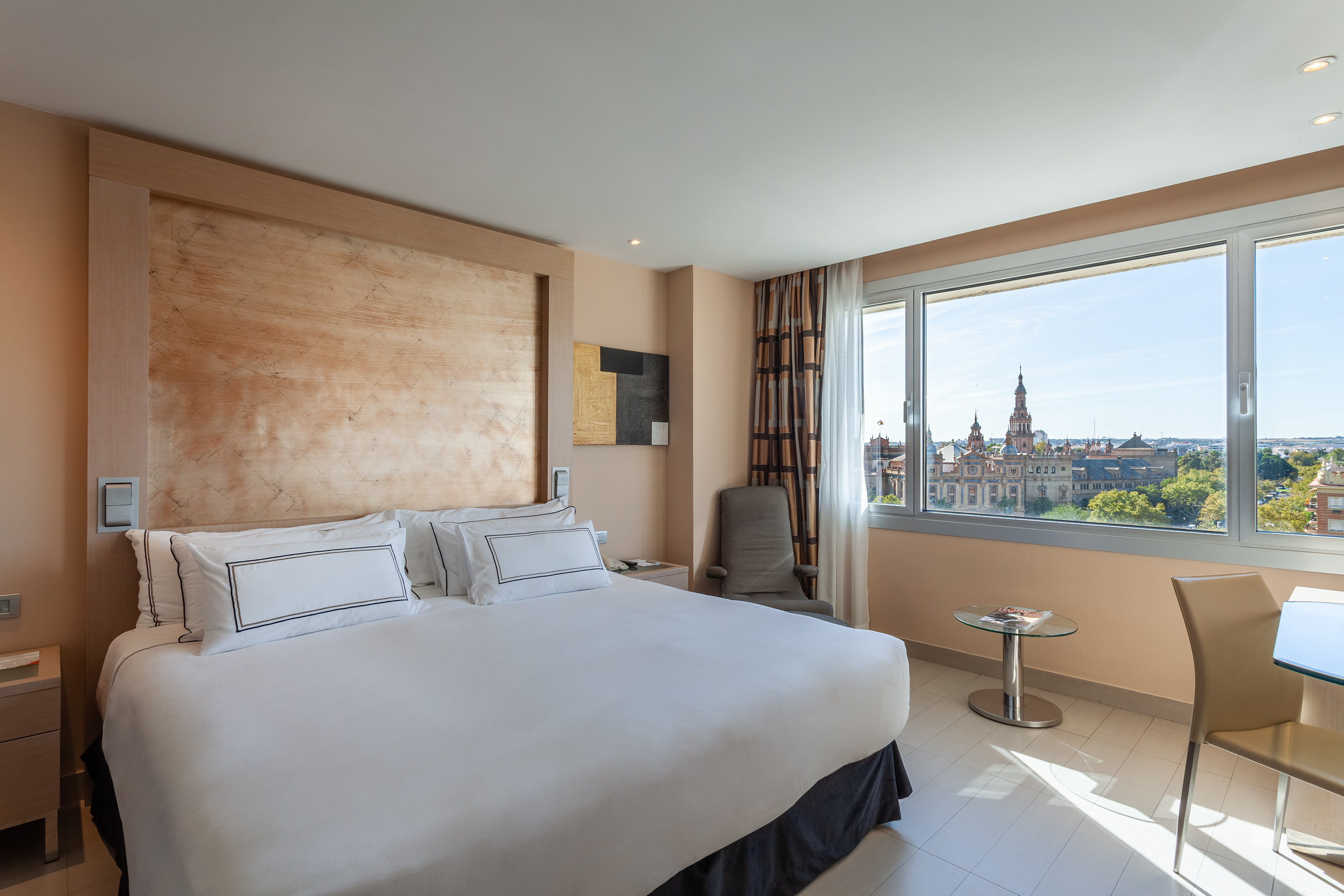 Hotel bedroom with large white bed, desk, and panoramic city view.