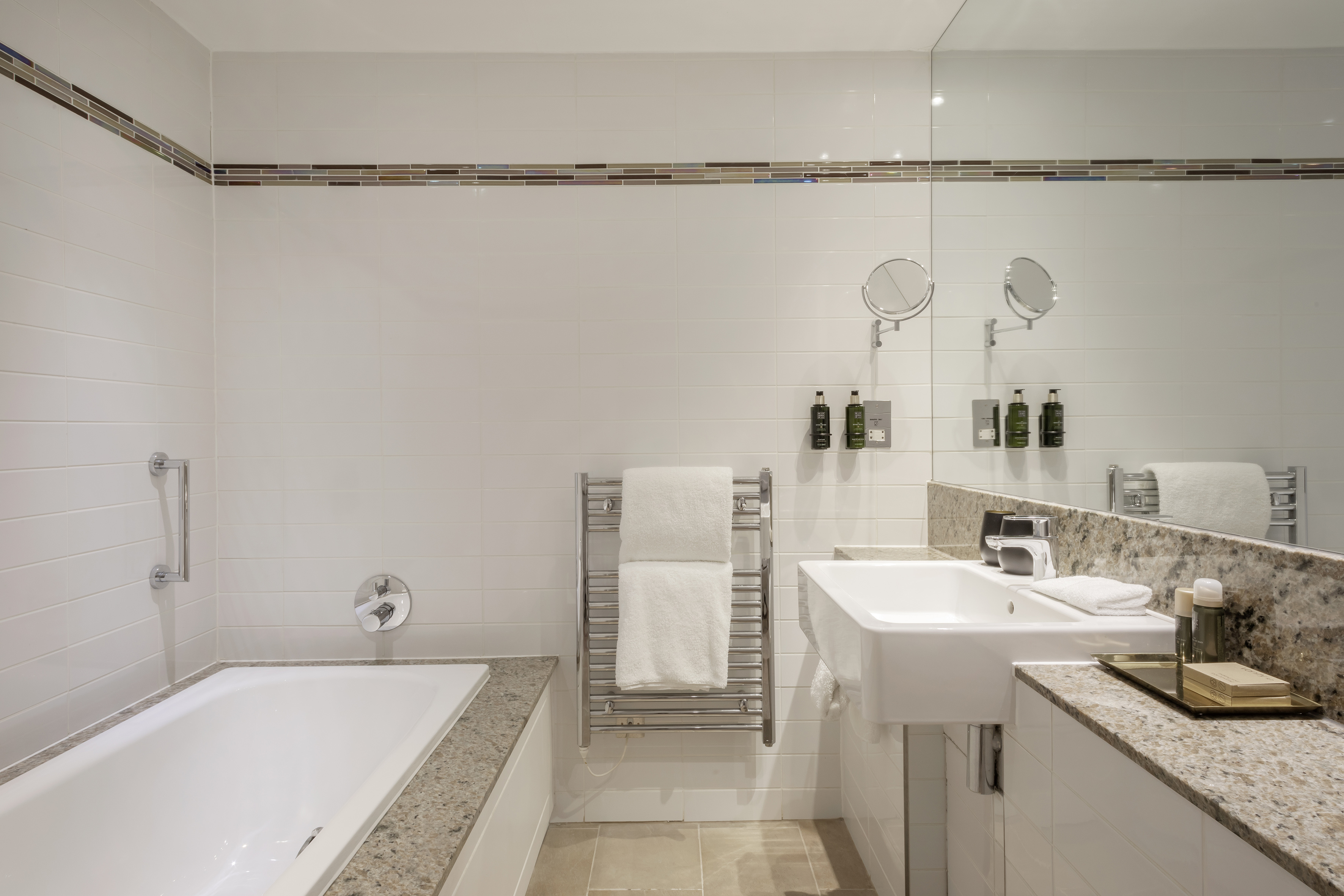 Modern hotel bathroom with white tiles, bathtub, sink, mirror, towel rail.