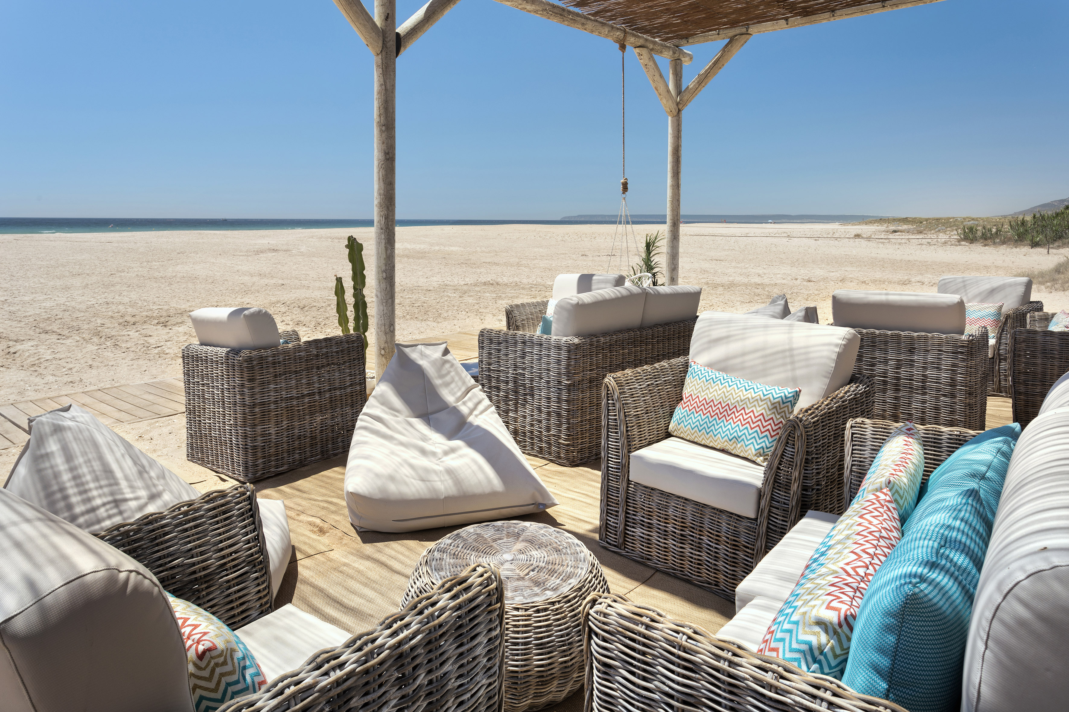 Wicker beach lounge with colorful pillows on wooden deck overlooking sandy ocean beach.
