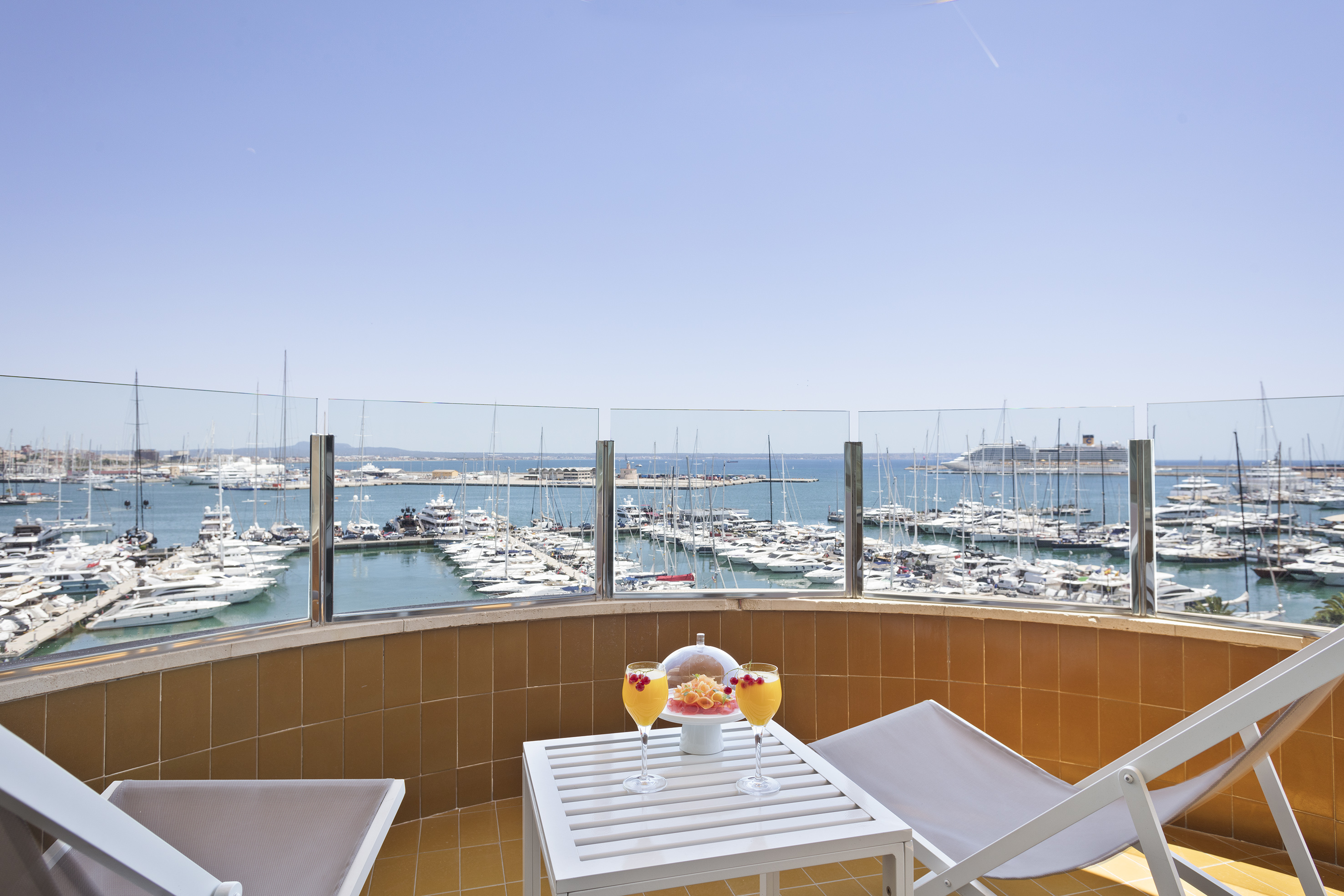 Sunny balcony with cocktails and lounge chairs, overlooking a yacht marina.