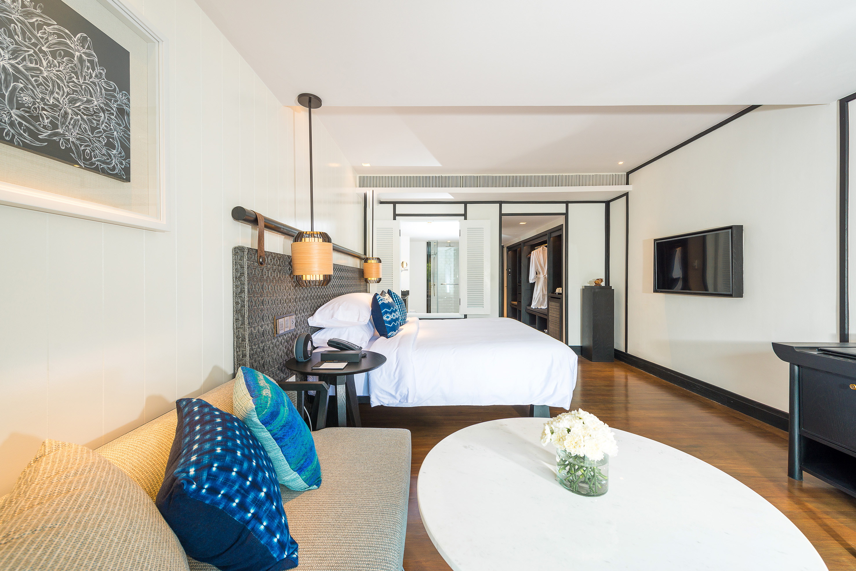 Modern hotel suite with white bed, blue pillows, sofa, TV, and hardwood floors.