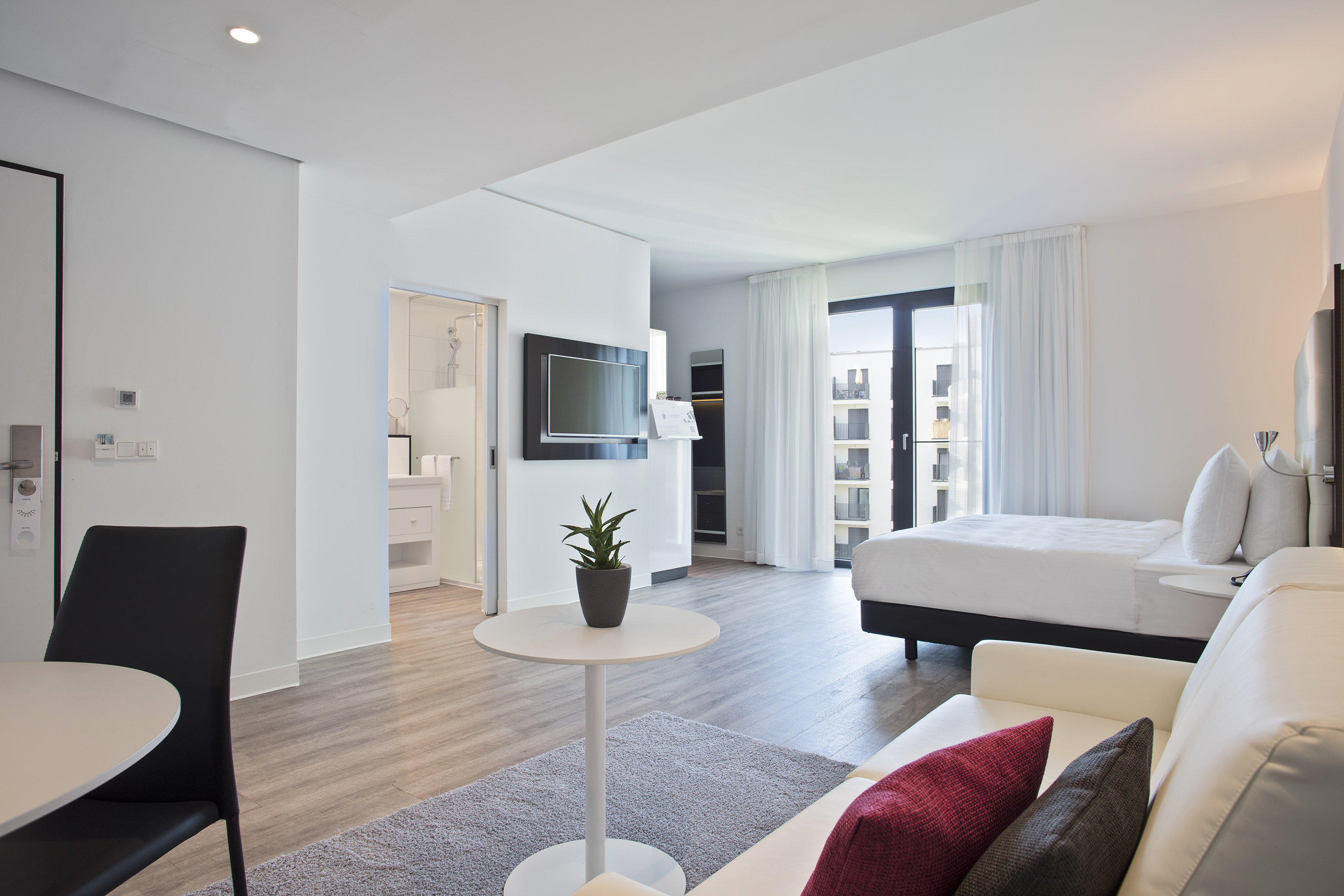 Bright modern hotel studio apartment with bed, sofa, TV, and large windows.