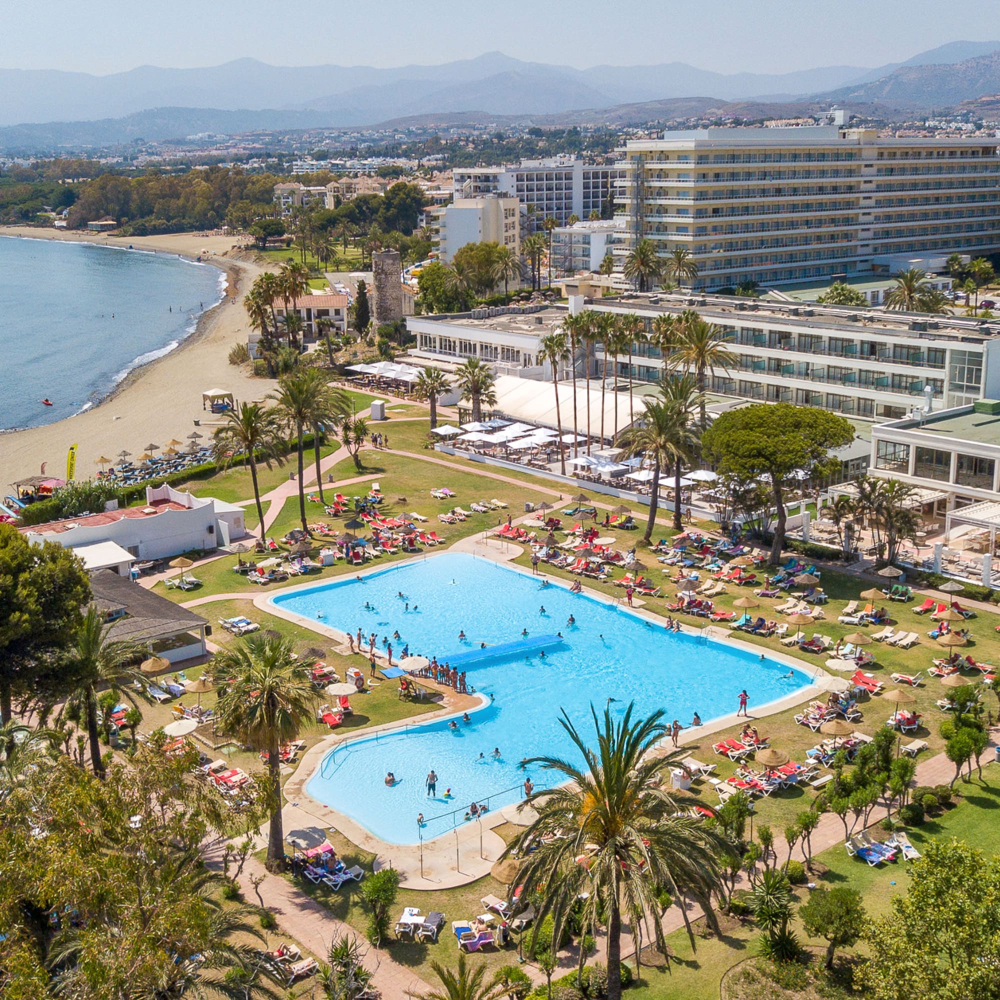 Beachfront hotel resort with large pool, sandy beach, and mountains.