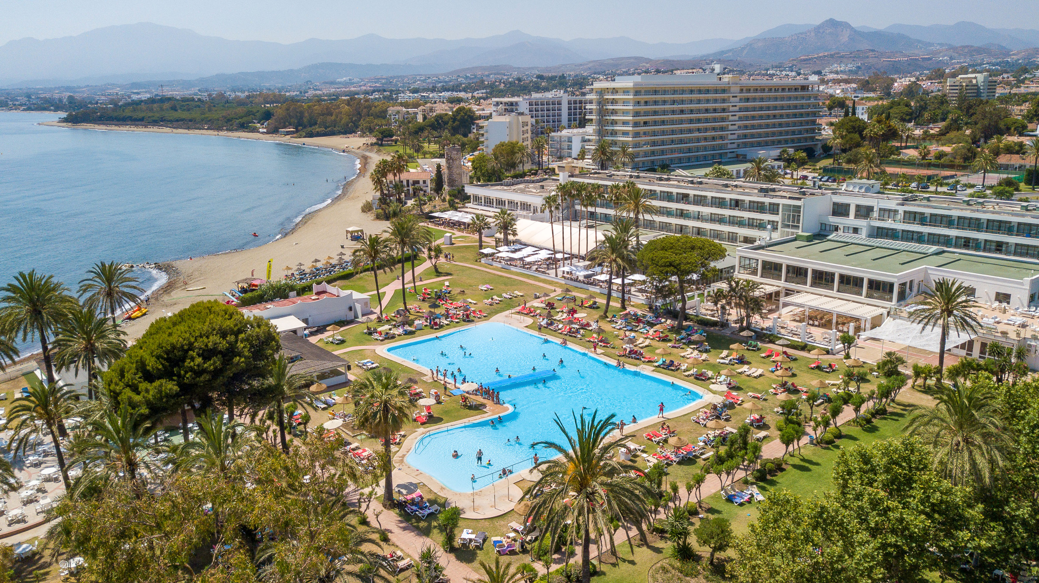 Beachfront hotel resort with large pool, sandy beach, and mountains.