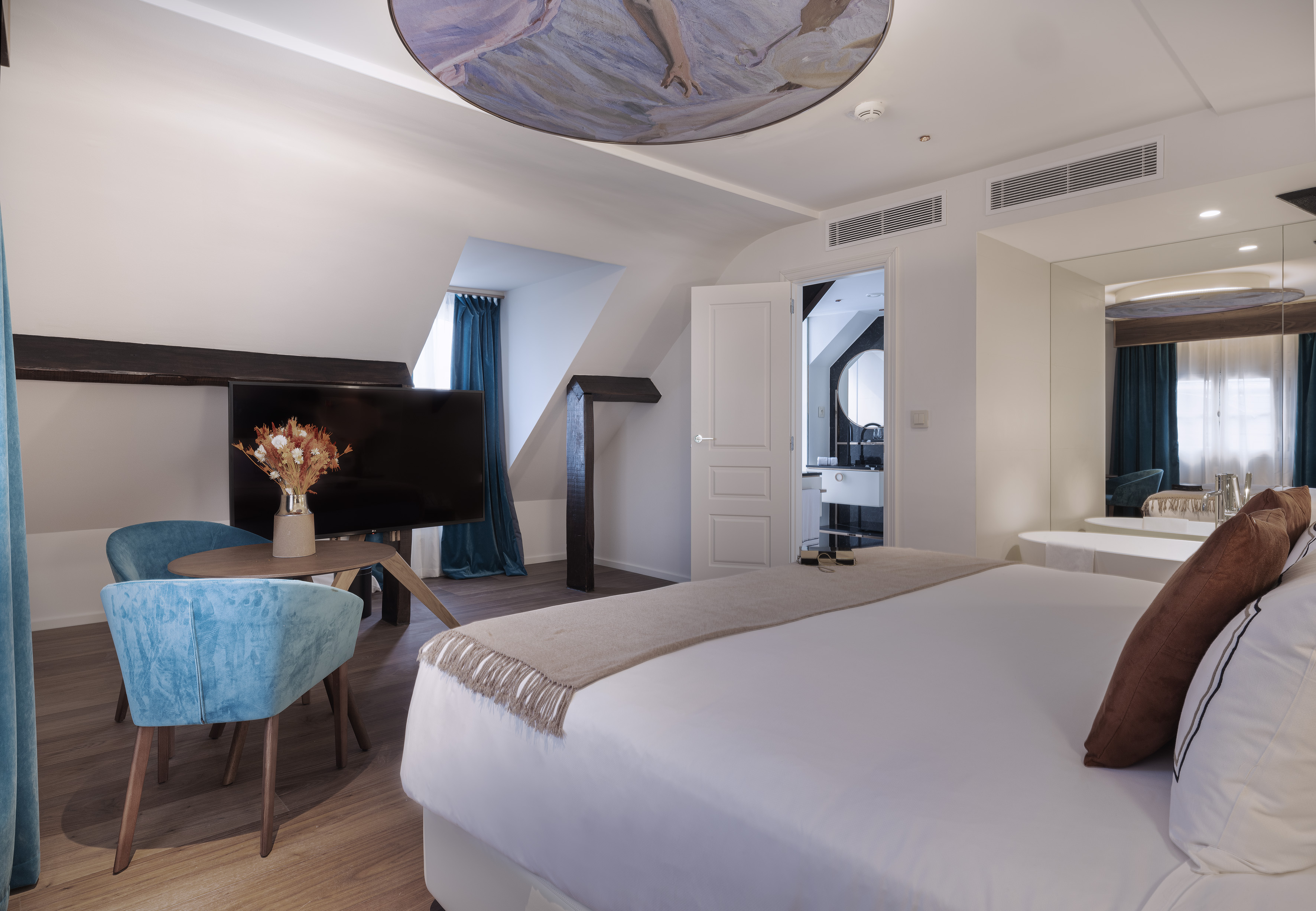 Modern attic hotel room with king bed, blue velvet chairs, ceiling mural.