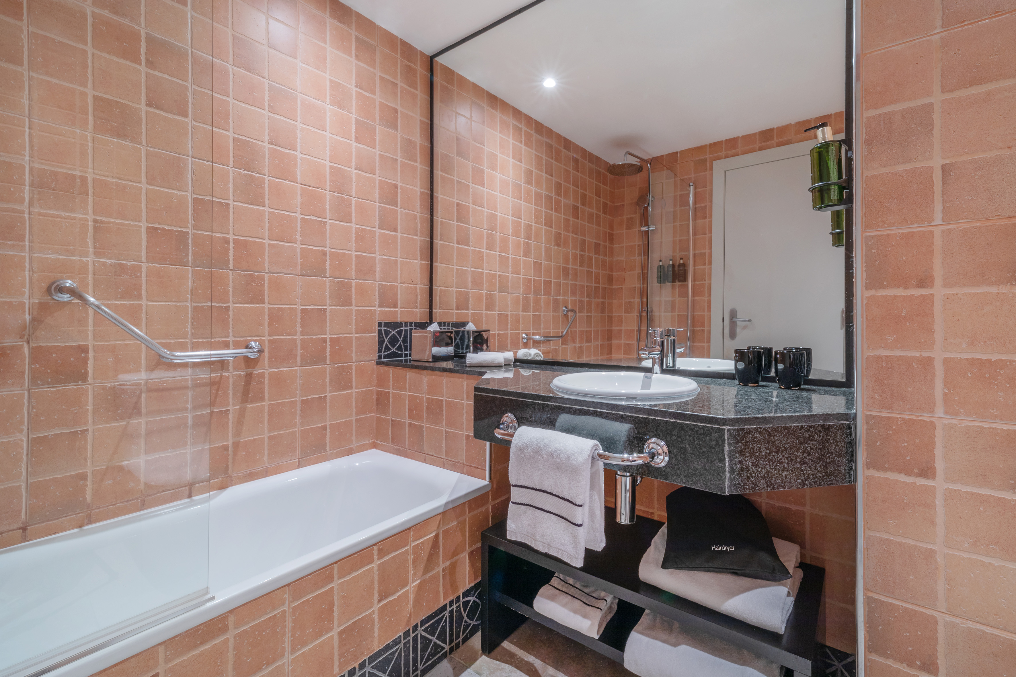 Hotel bathroom with warm terracotta tiles, bathtub, sink vanity, and large mirror.