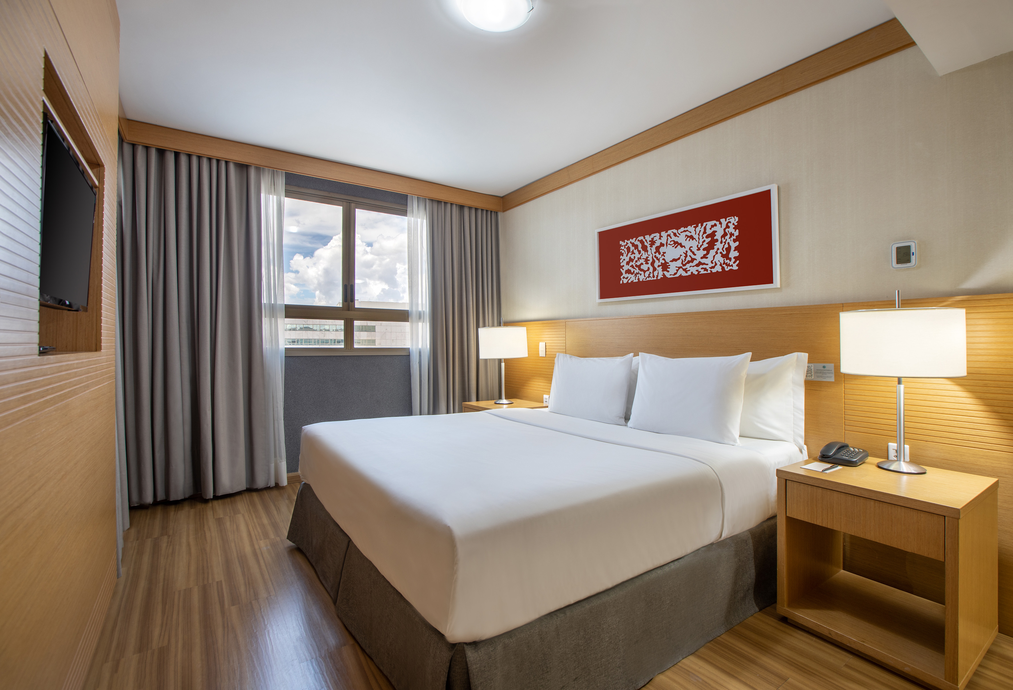 Modern hotel room with king bed, white bedding, wood floor, and large window.
