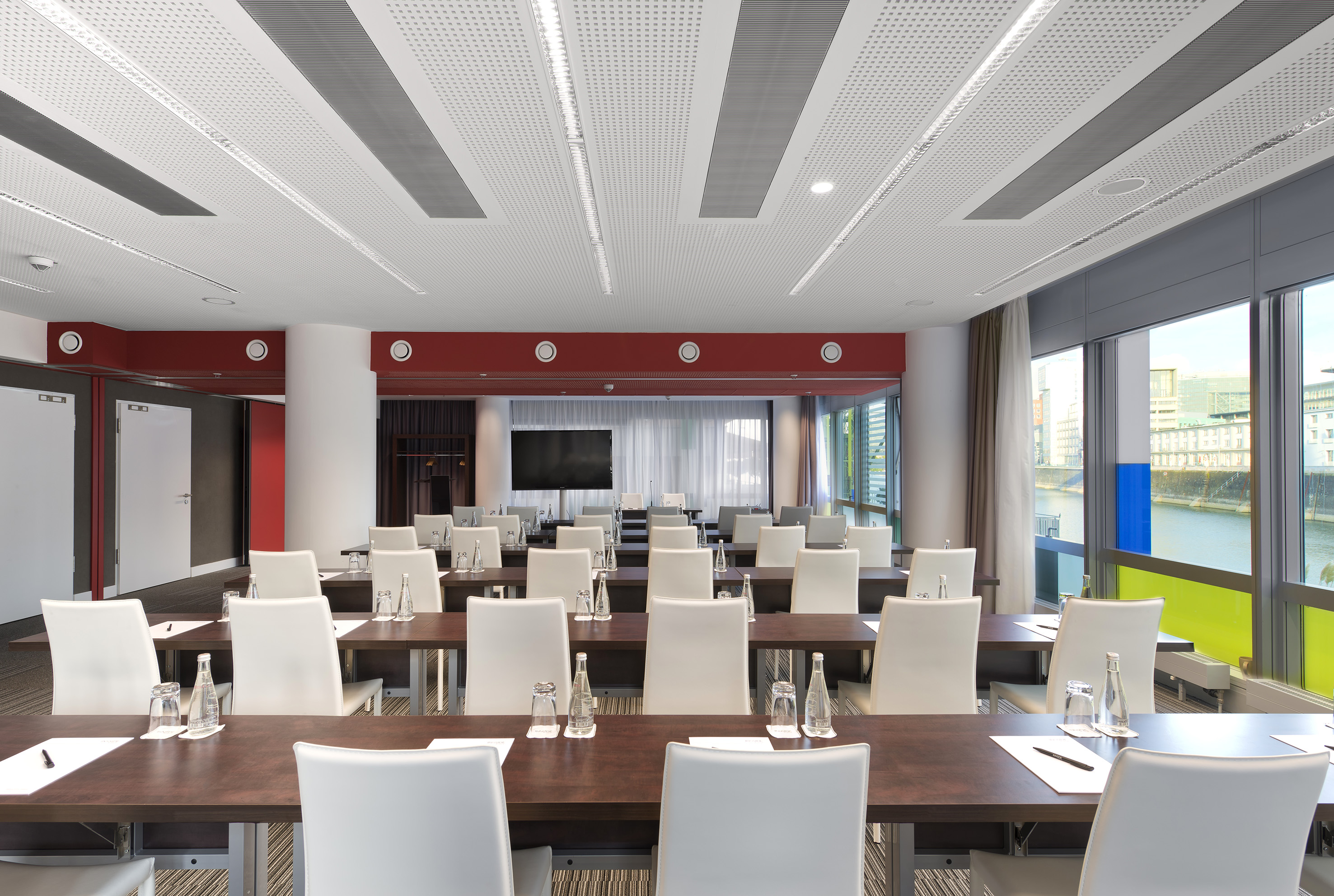 Modern conference room with rows of tables, white chairs, large screen, and river view.