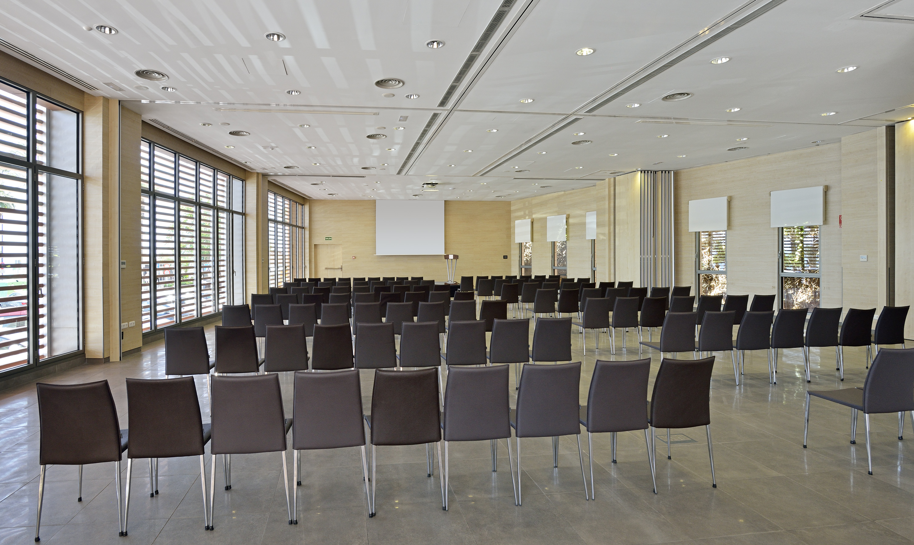 Empty modern conference room with dark chairs, projection screen, windows.