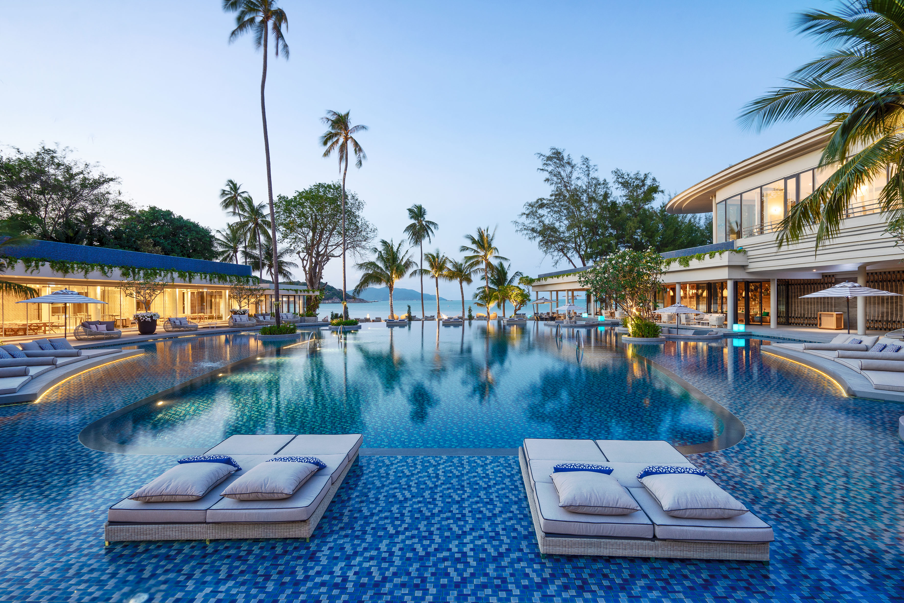 Luxury resort pool with palm trees, ocean background, and lounge chairs at dusk.