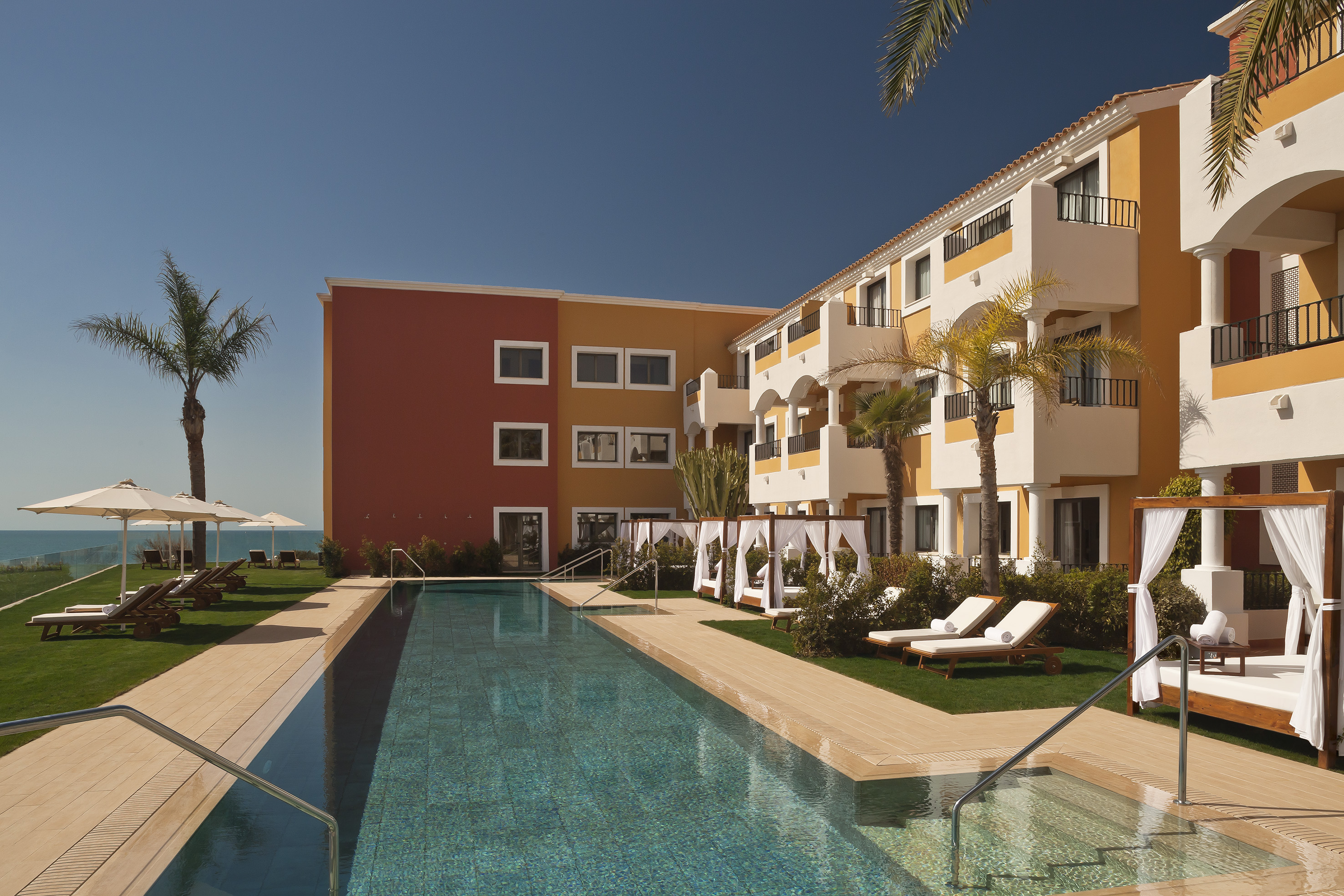 Resort swimming pool with colorful hotel, palm trees, loungers, and sea view.