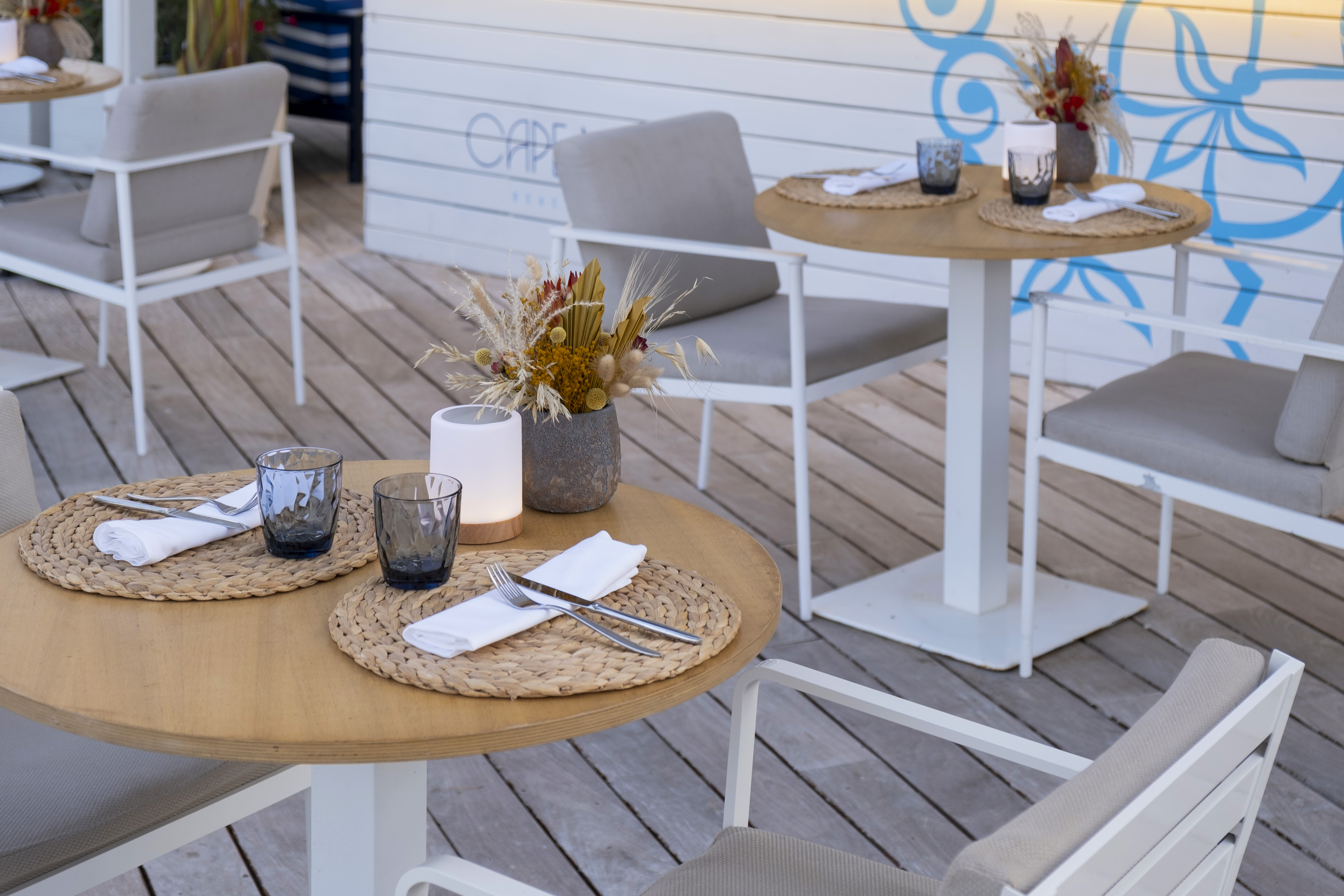 Outdoor restaurant patio with set tables, white chairs, and dried flower decor.