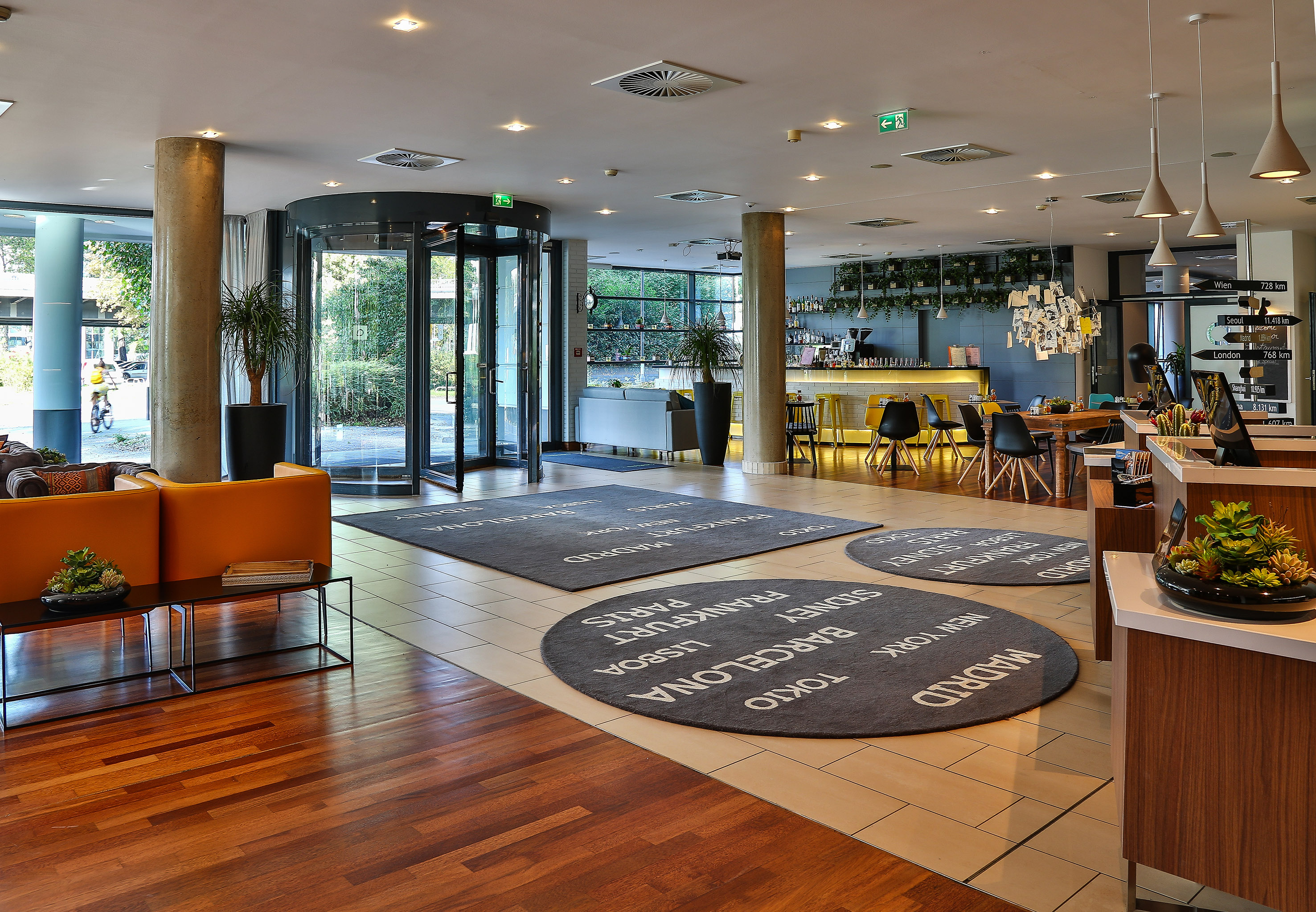 Modern hotel lobby with revolving door, bar, and city name rugs on the floor.