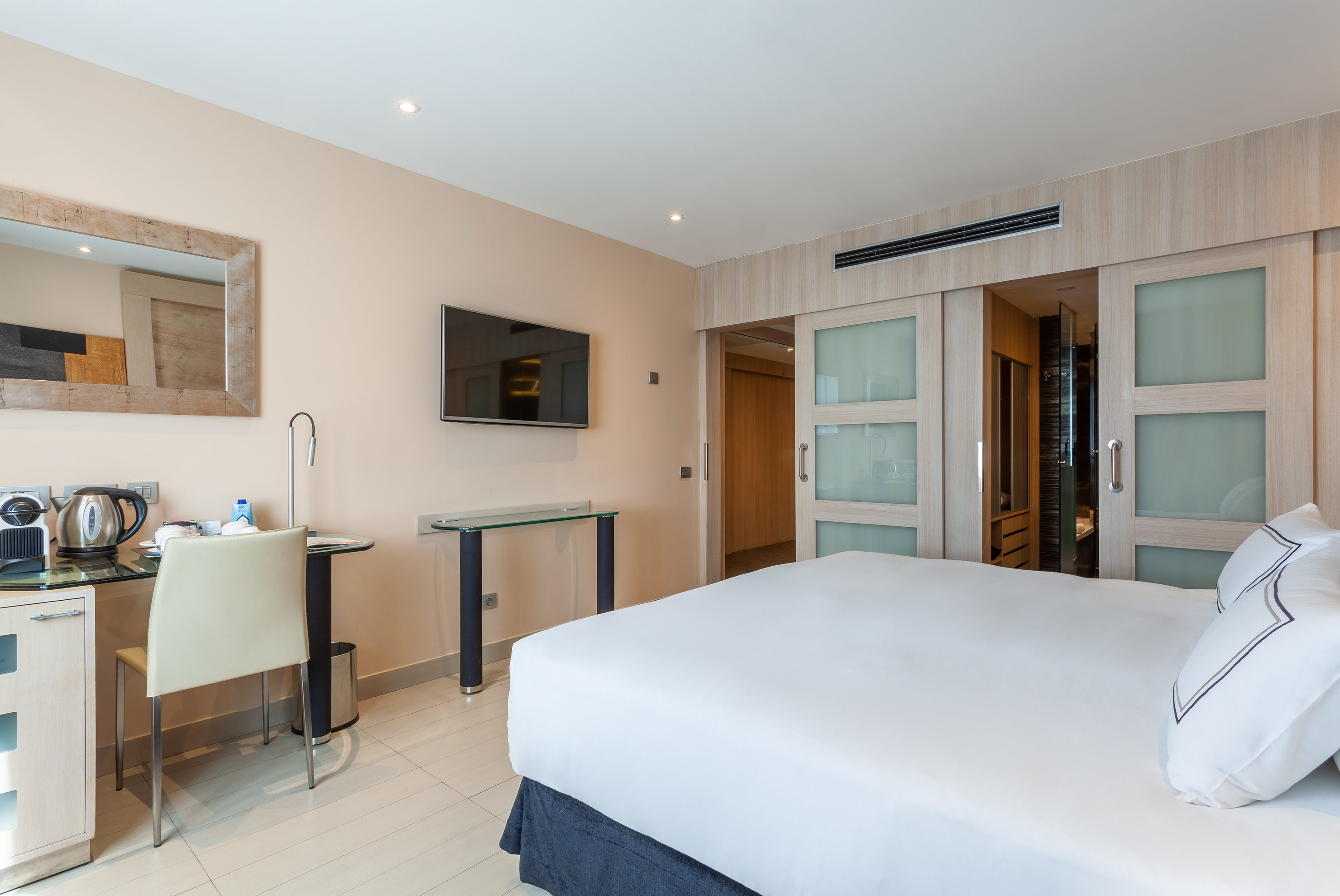 Modern hotel bedroom with white bed, mounted TV, desk, and sliding wardrobe.