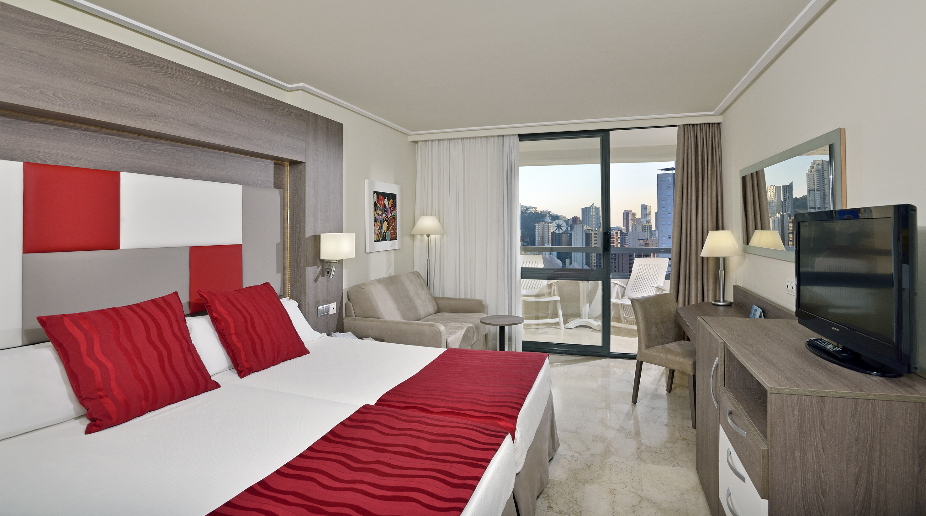 Modern hotel room with twin beds, red accents, desk, and balcony city view.