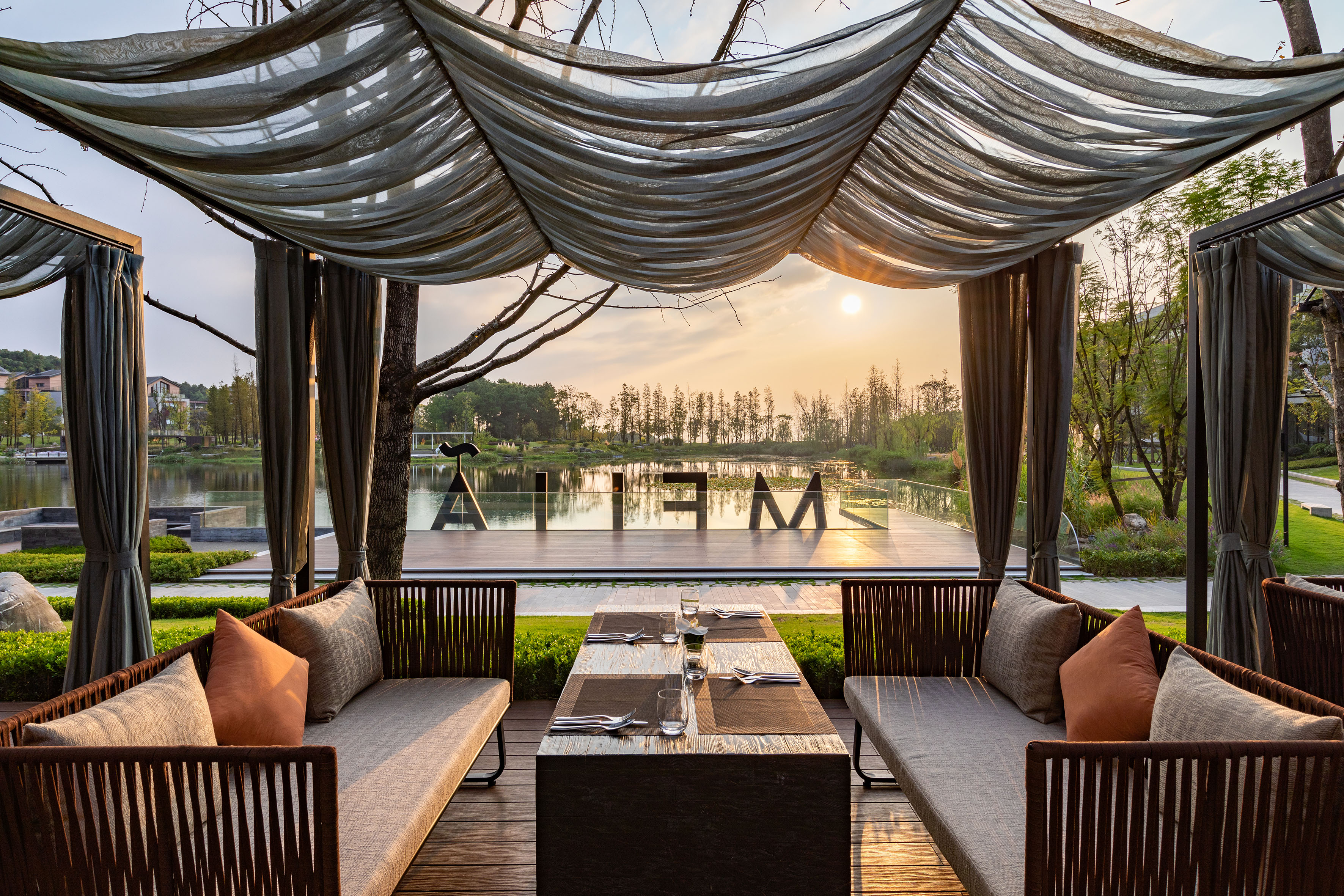 Lakeside outdoor dining patio at sunset, elegant table setting under draped canopy.