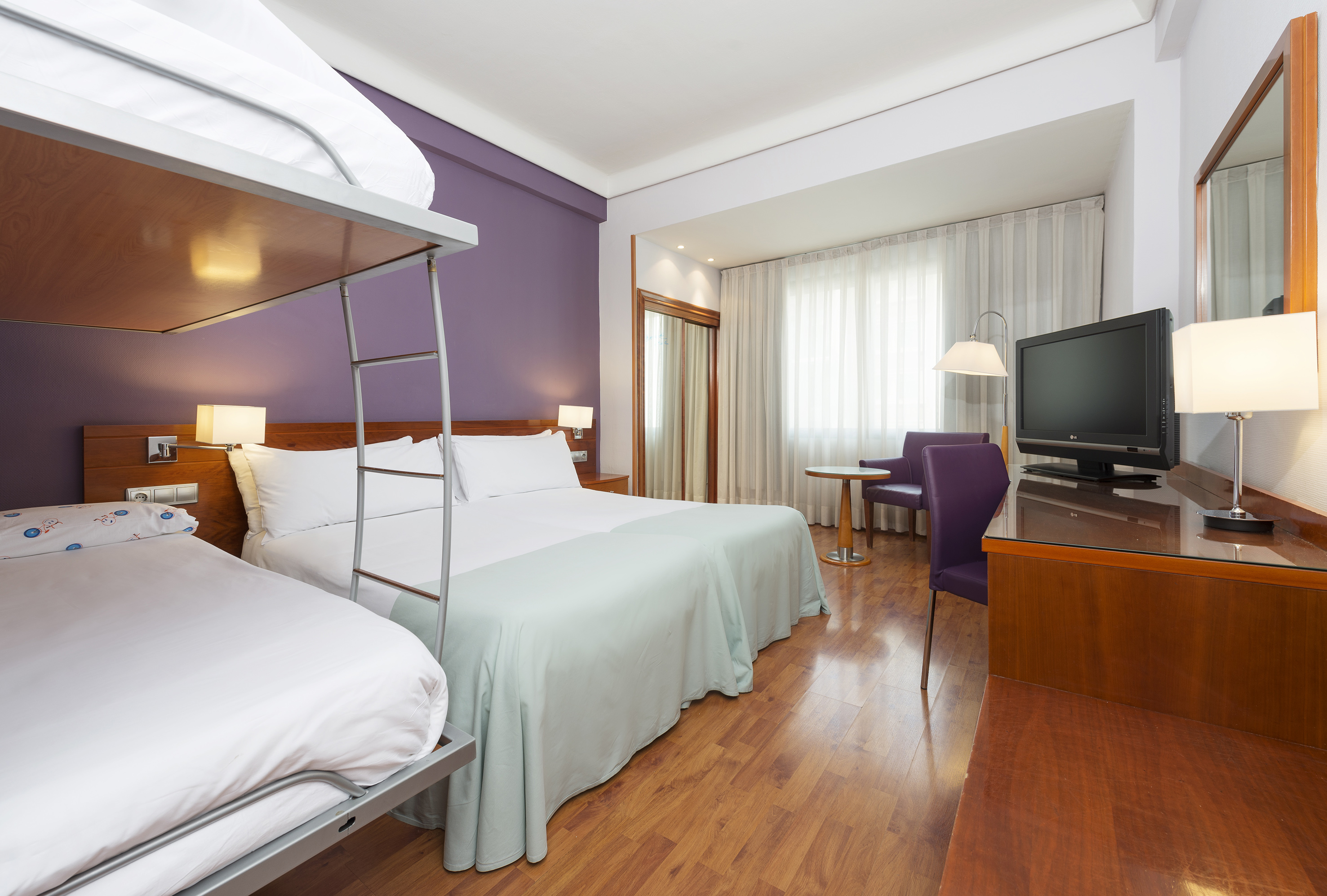 Hotel room: bunk bed, large bed, purple wall, desk with TV, wood floor.