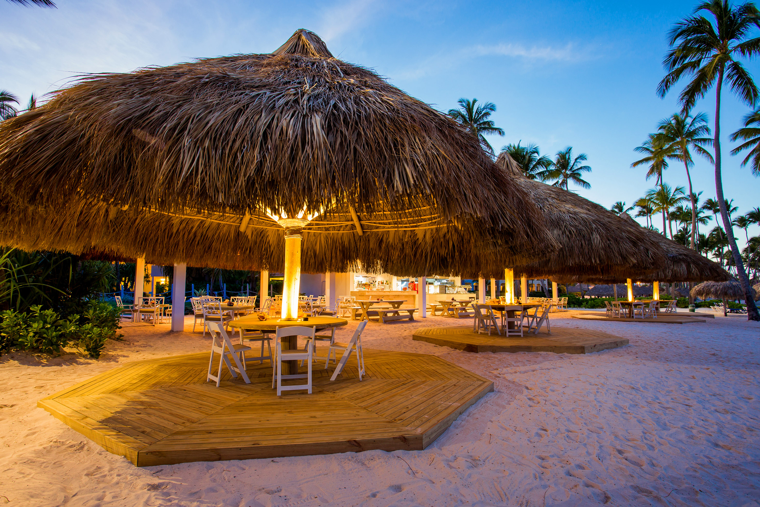 Tropical beach bar with illuminated thatched roofs and outdoor dining at dusk.