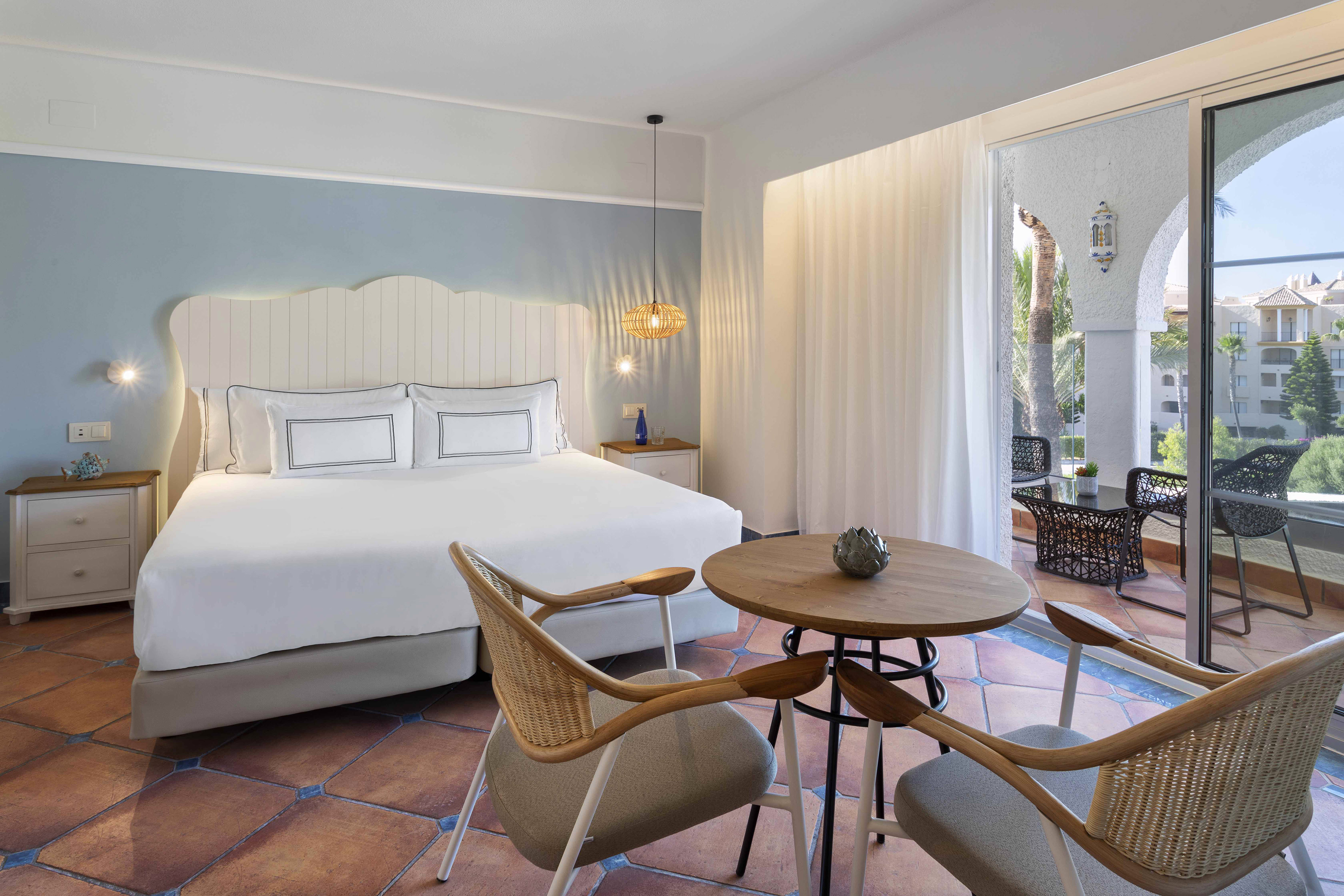Coastal hotel bedroom with white bed, rattan chairs, terracotta floor, and sunny balcony.