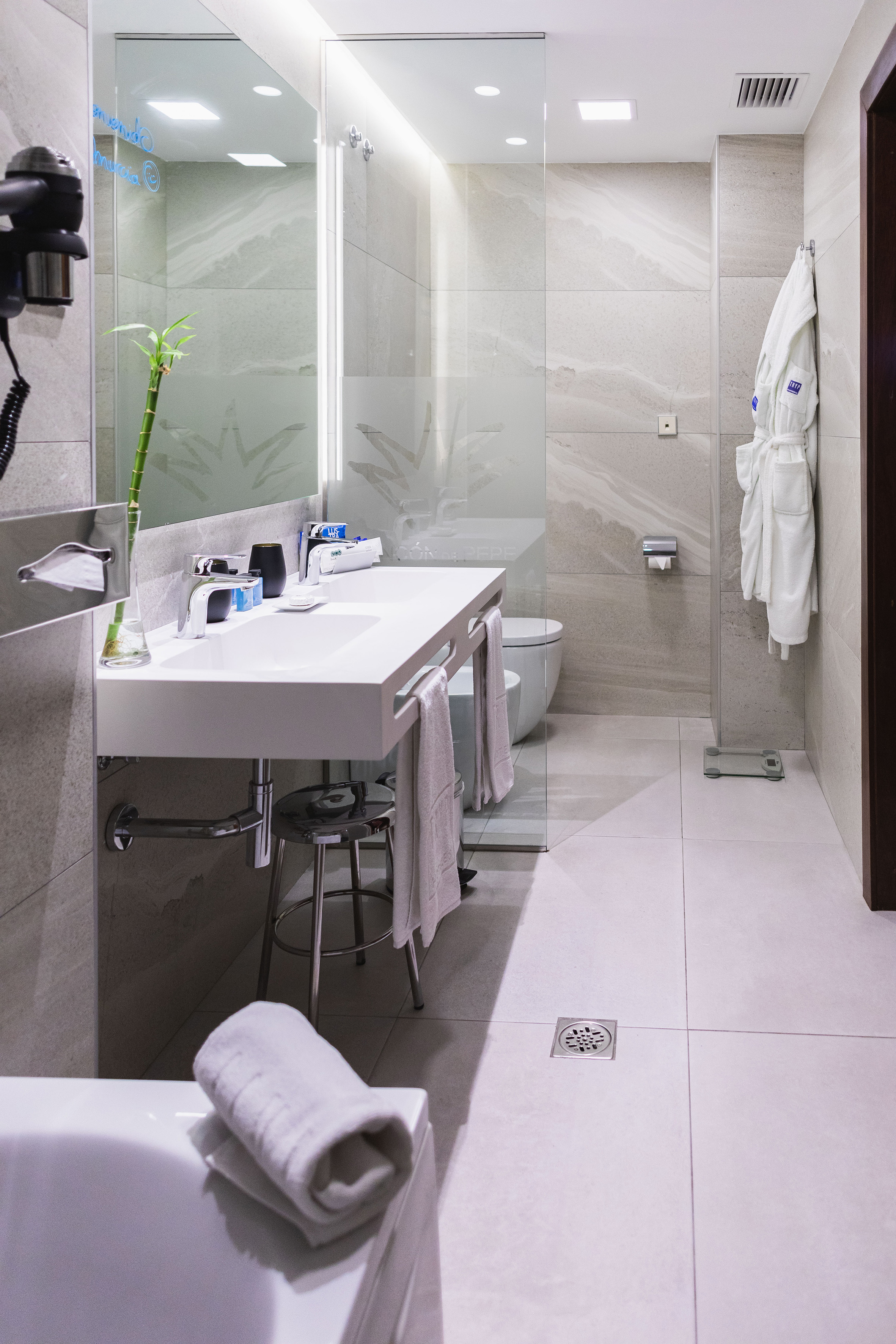 Modern hotel bathroom with white vanity, grey tiles, glass shower, toilet, and bathrobe.