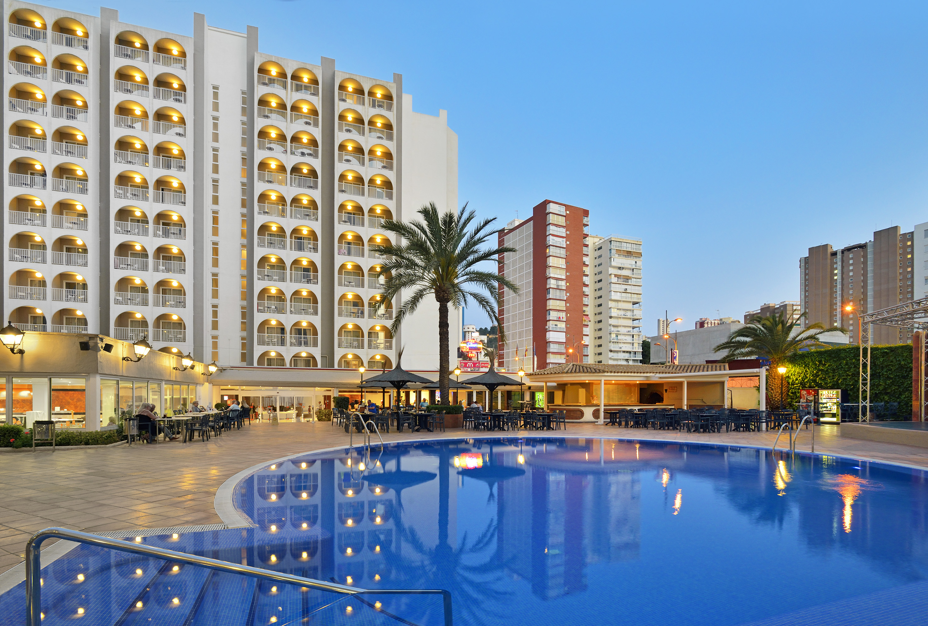 Evening hotel pool with illuminated balconies, outdoor dining, and palm trees.