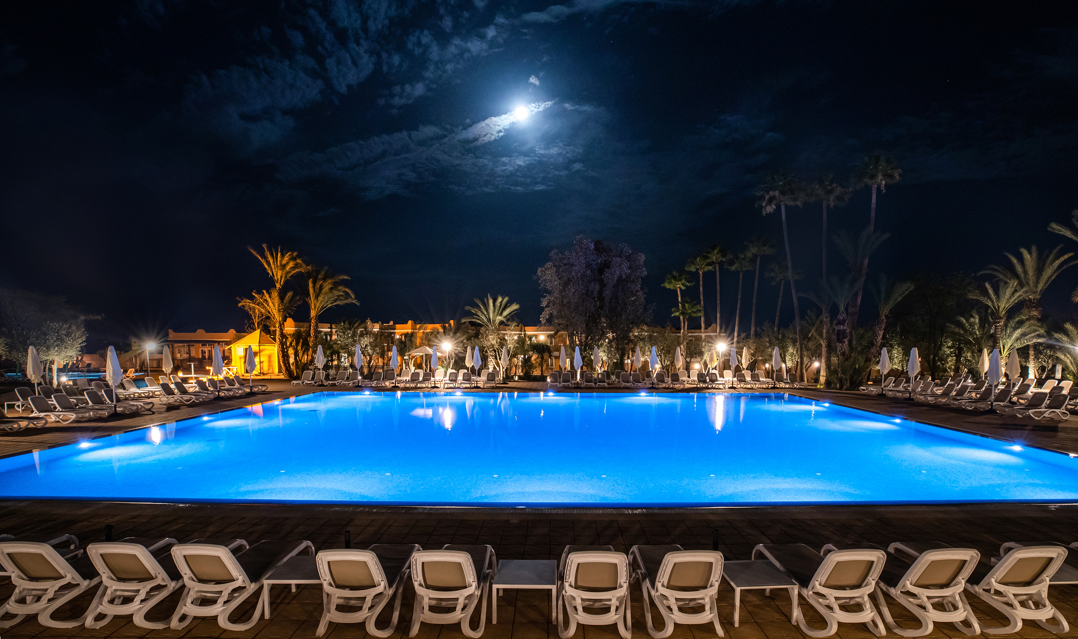 Moonlit resort pool at night with glowing blue water and palm trees.