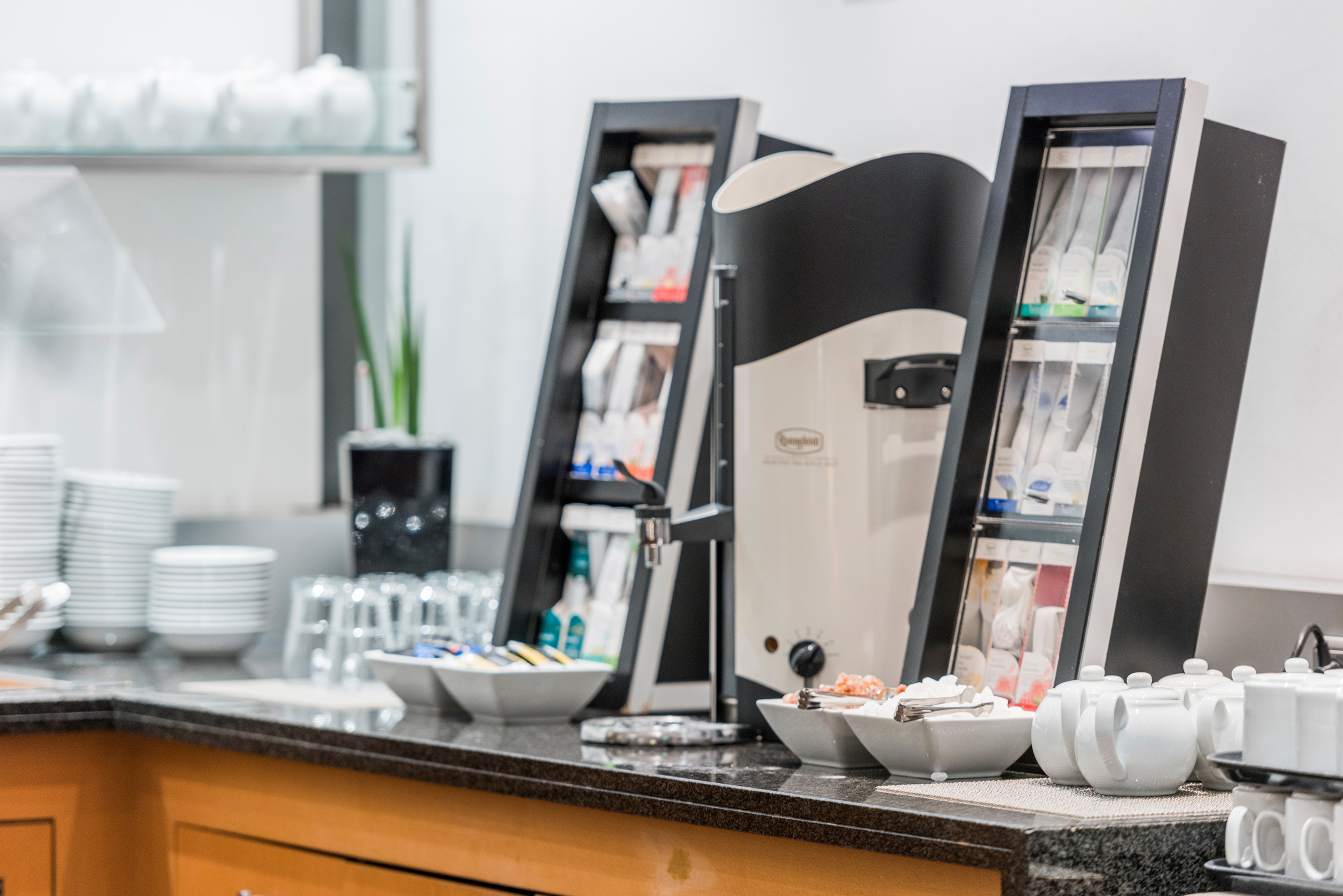 Self-service tea and coffee station with hot water dispenser, tea bags, cups.