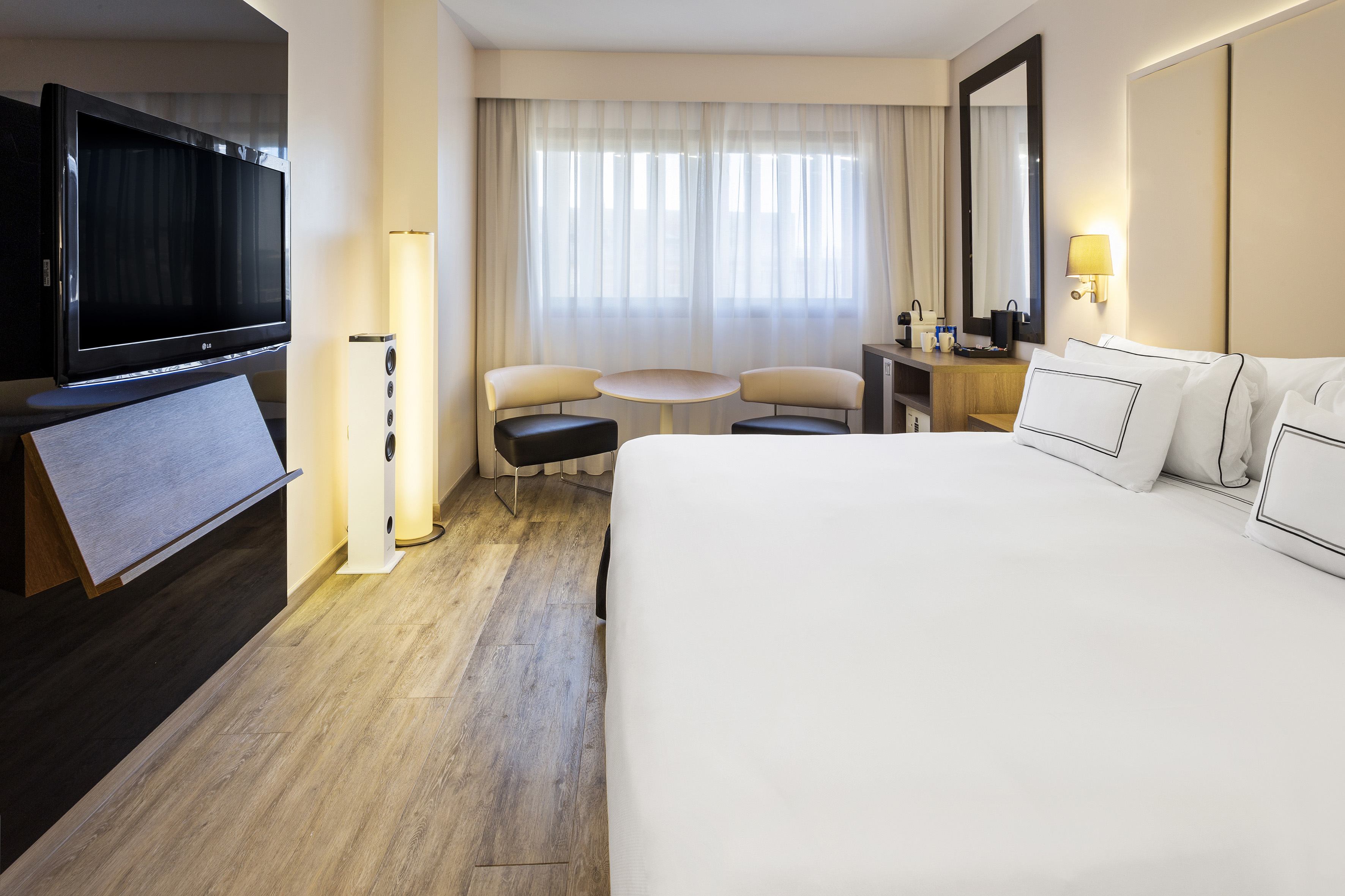 Modern hotel room with white king bed, flat-screen TV, wood floor, and sitting area.
