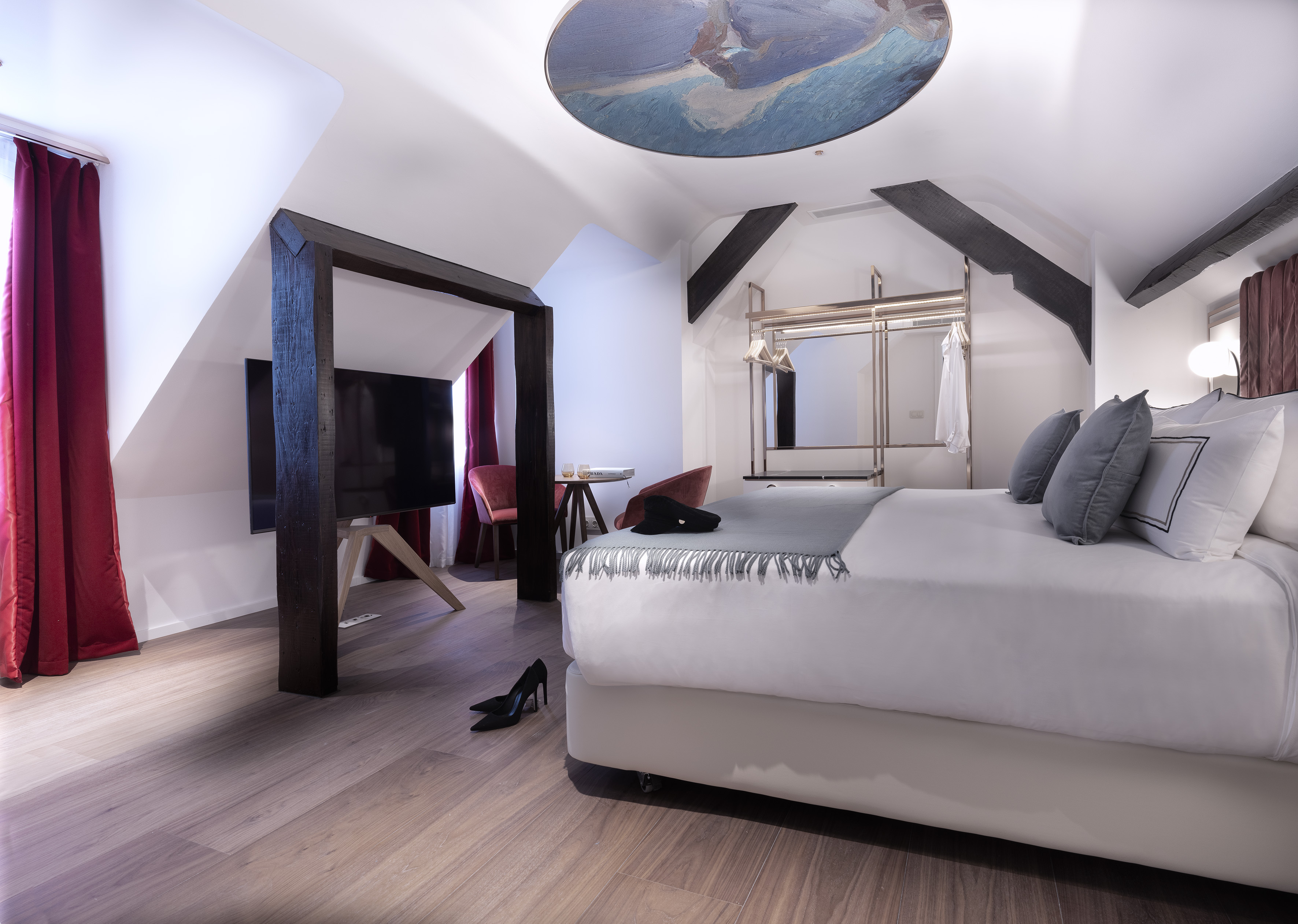 Attic bedroom with white bed, exposed wooden beams, ceiling mural, and red curtains.