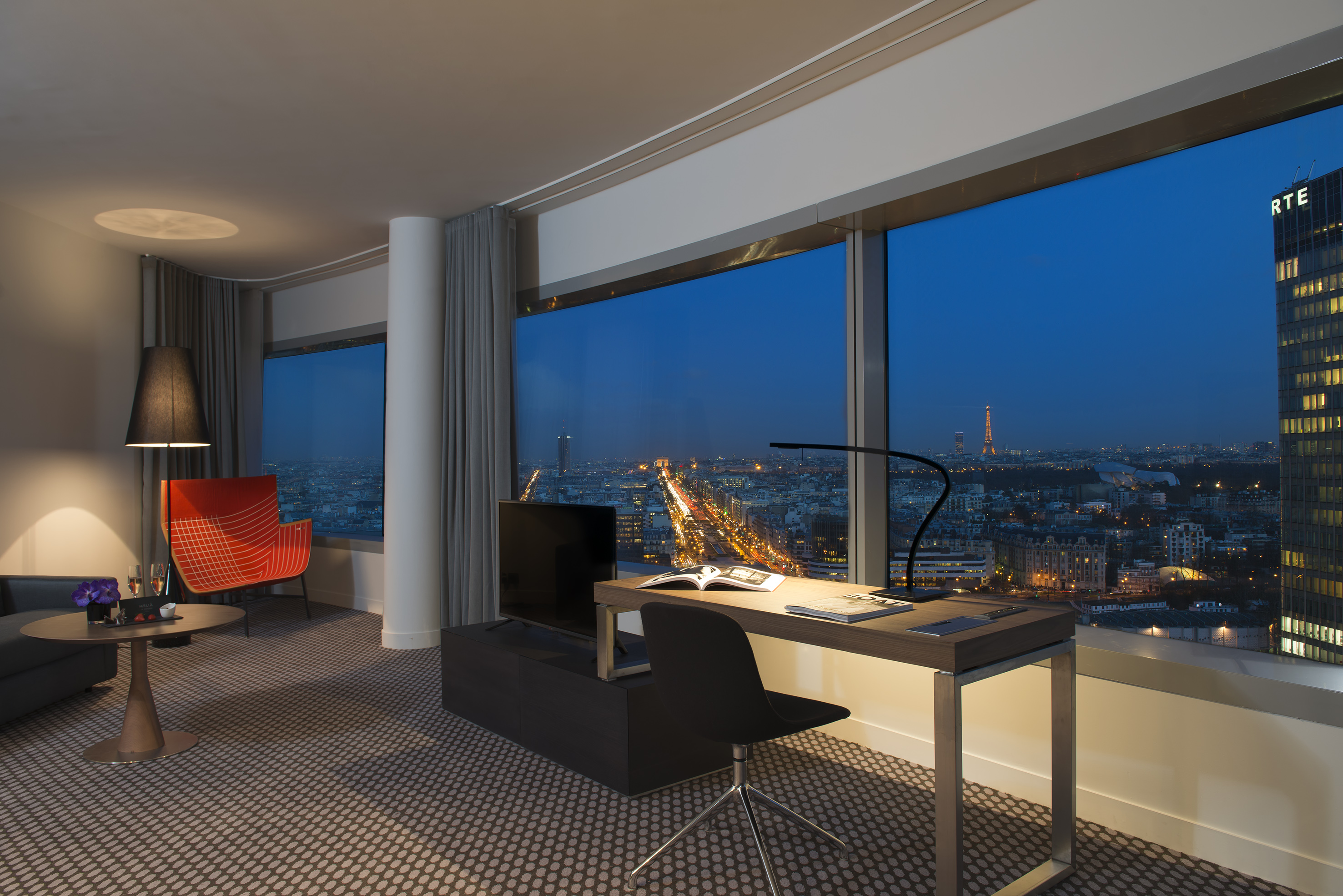 Modern hotel room with desk, panoramic Paris night view, Eiffel Tower visible.