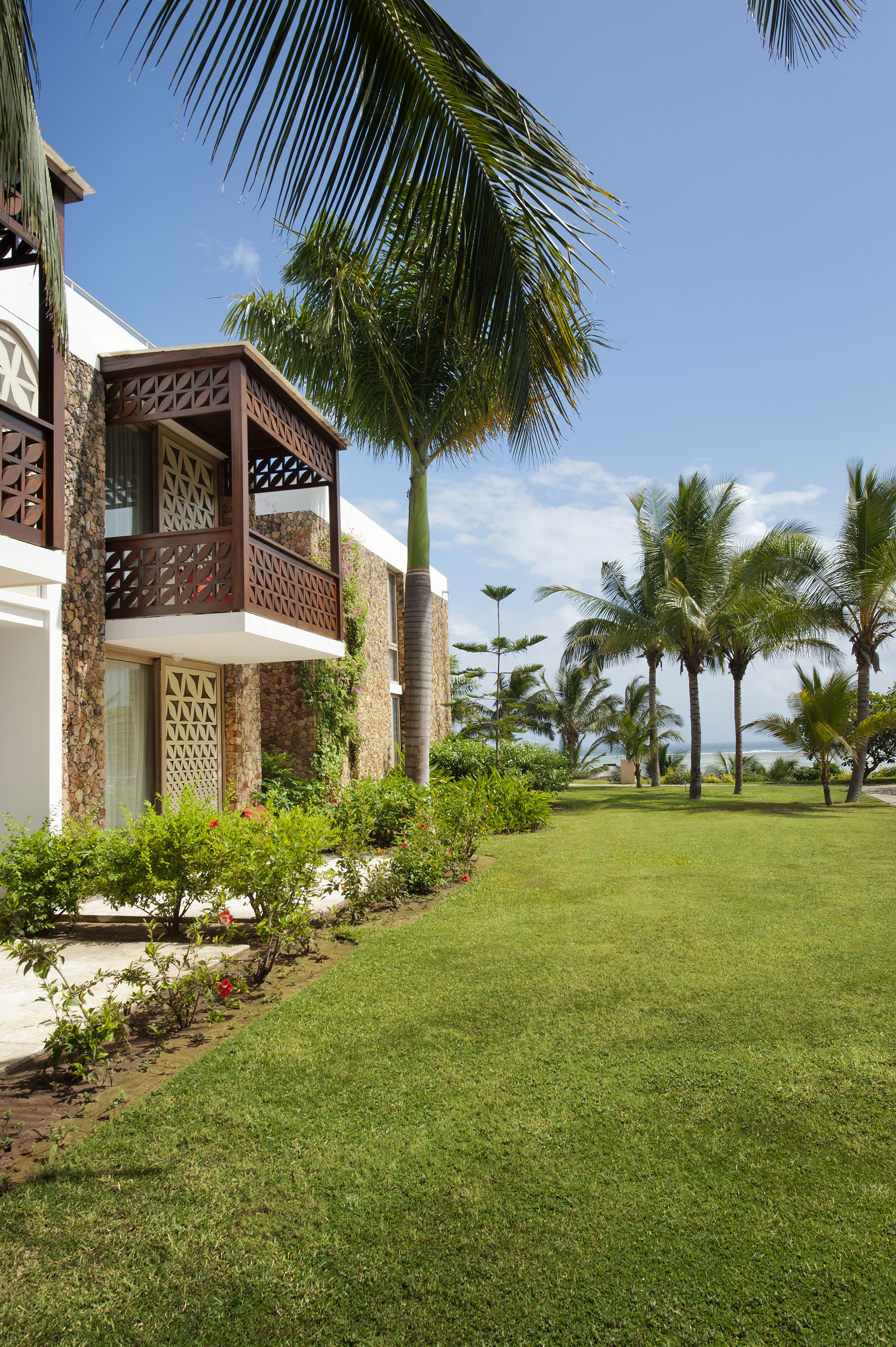 Tropical resort villa with ornate wooden balcony, green lawn, palm trees, and ocean view.