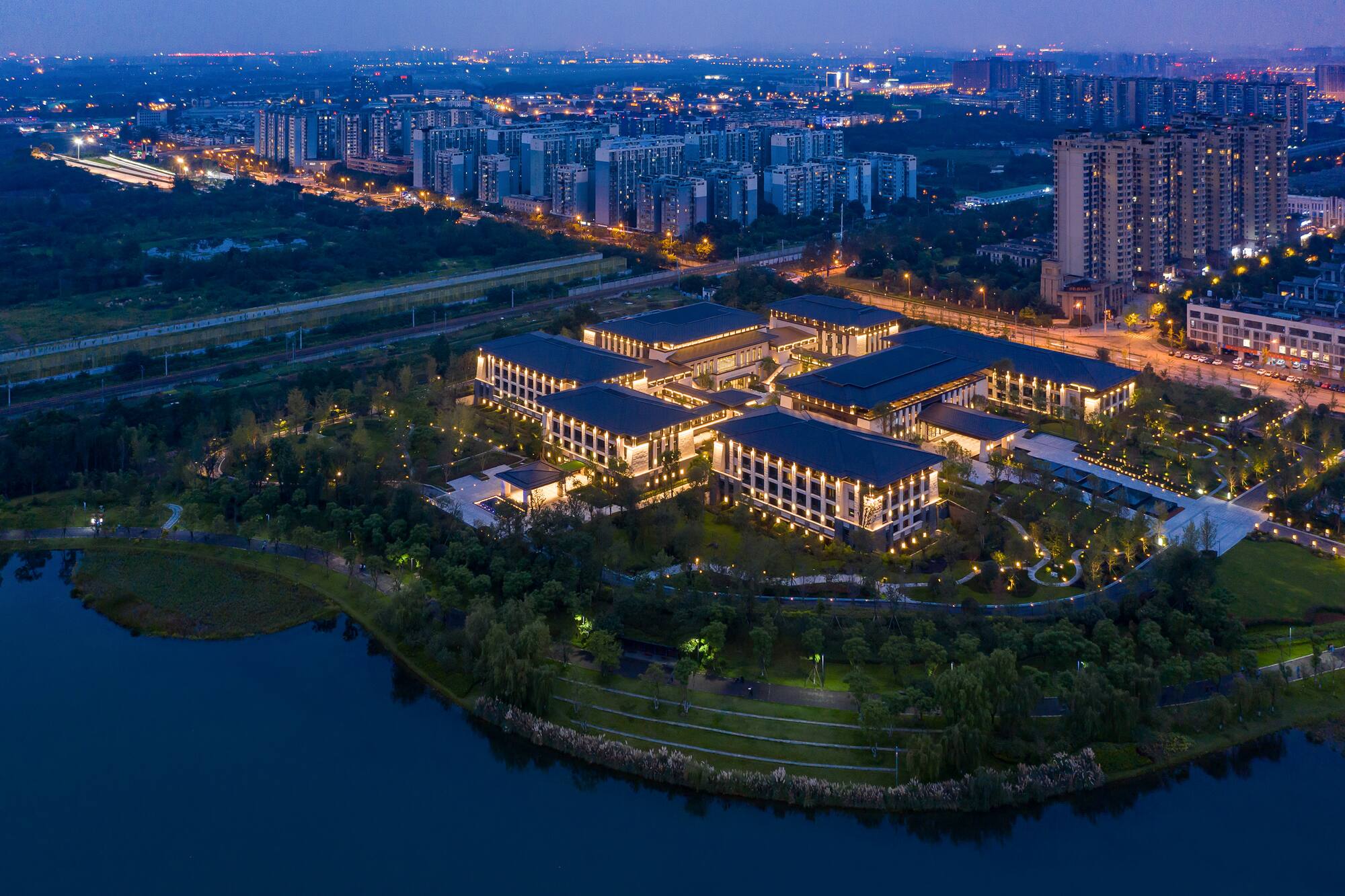 Aerial night view: Illuminated modern architecture by lake, with distant city skyline.