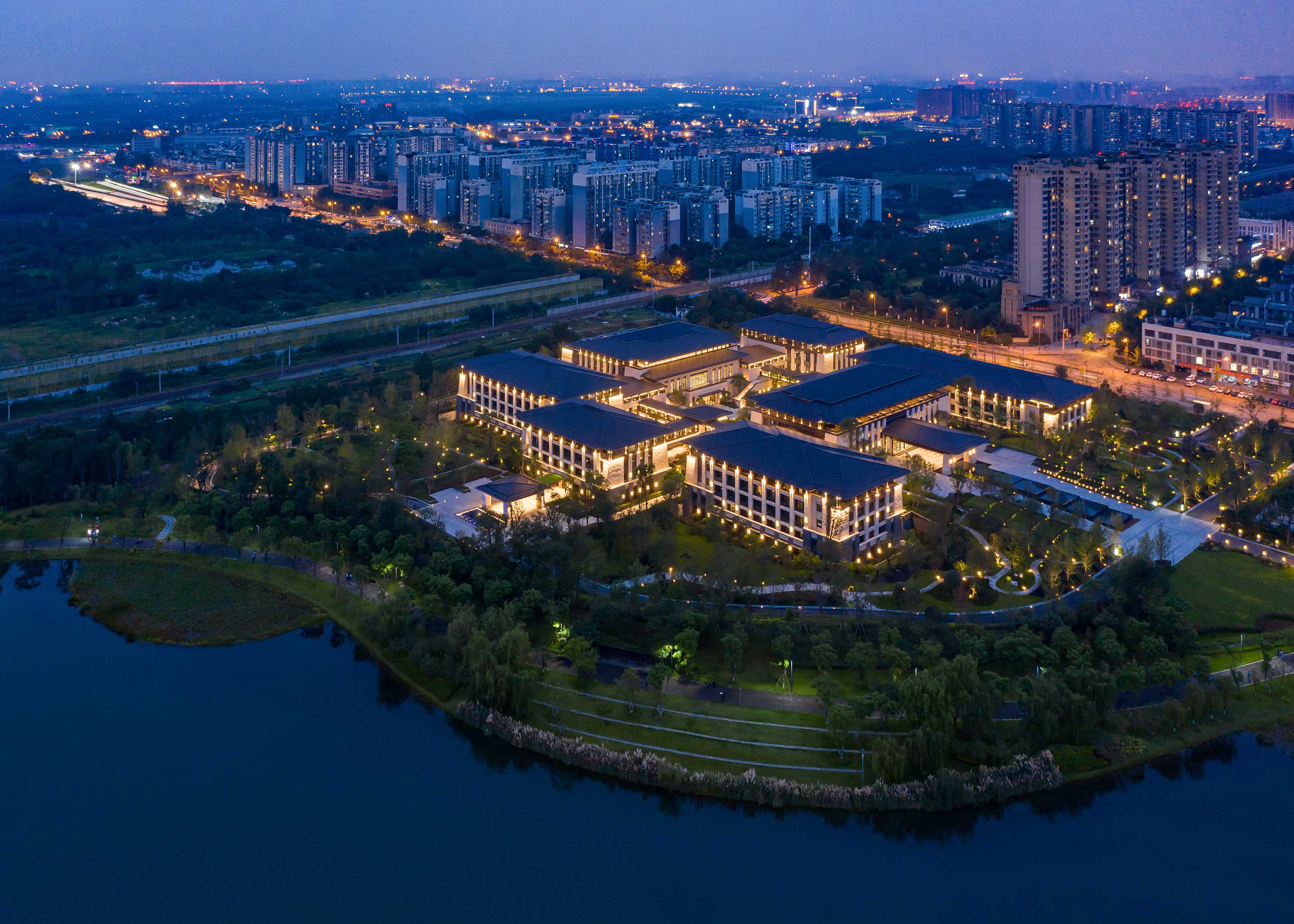 Aerial night view: Illuminated modern architecture by lake, with distant city skyline.
