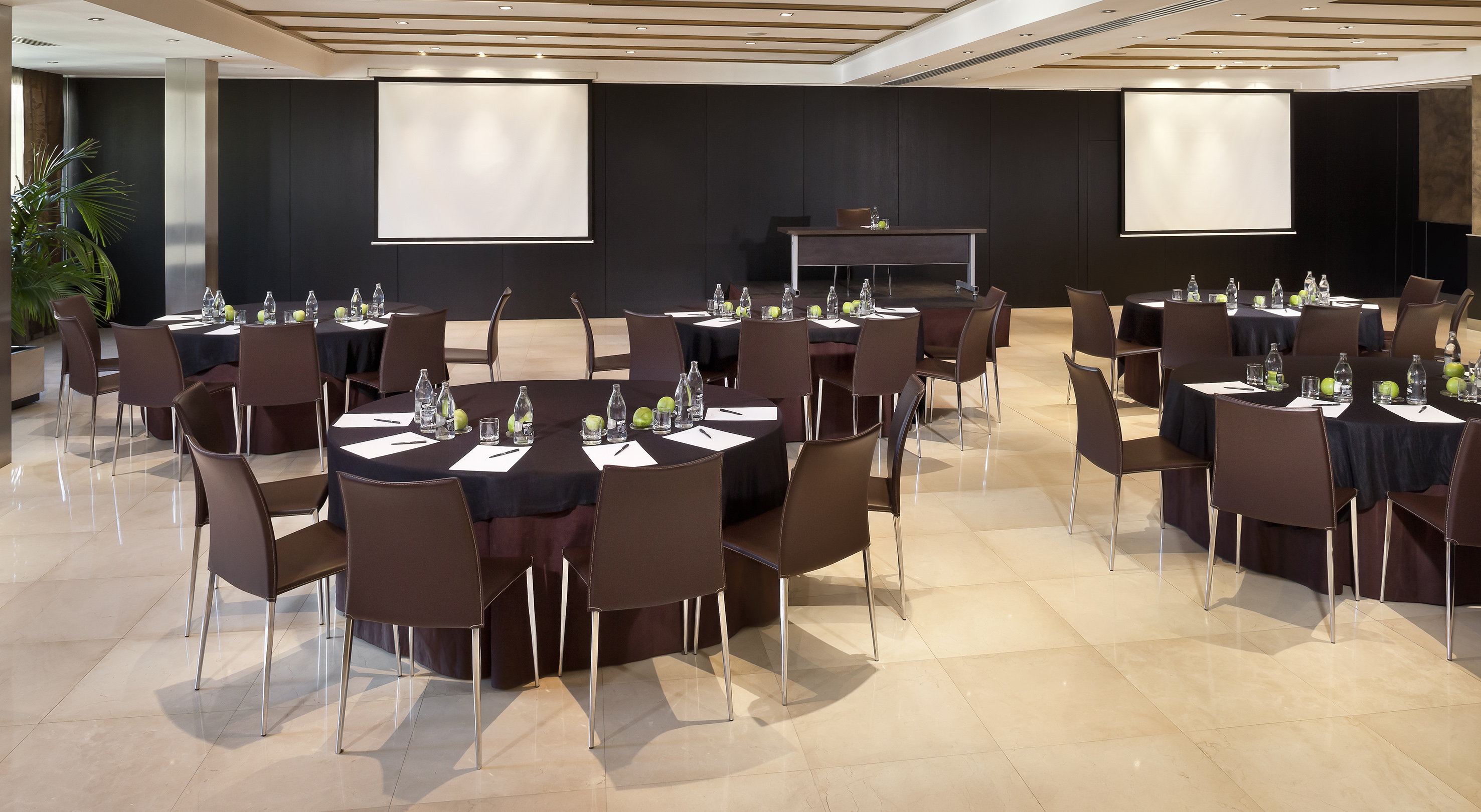 Modern conference room with round tables, brown chairs, refreshments, screens.